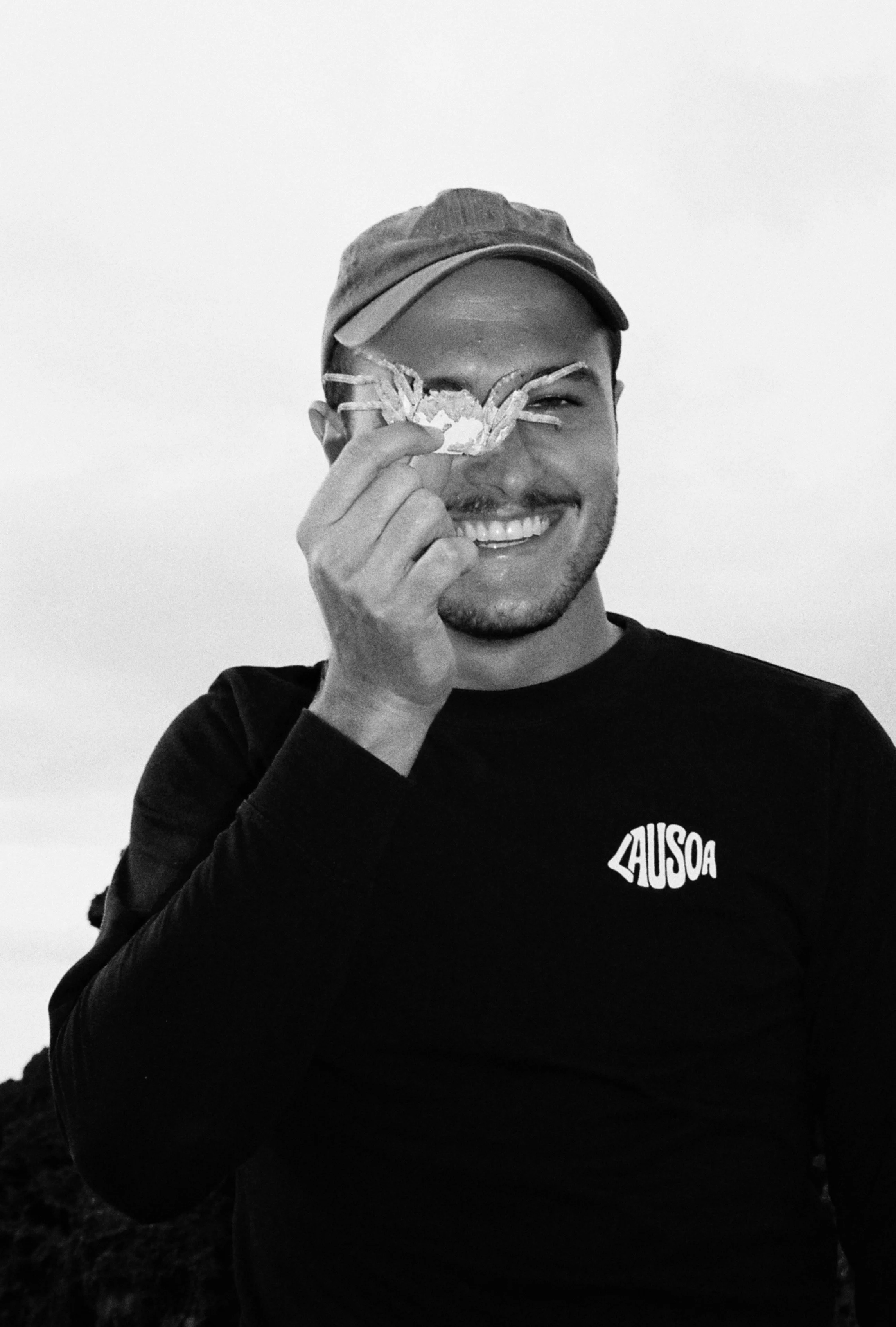 Un homme souriant tient une petite créature de crabe près de son œil. La photo est en noir et blanc.