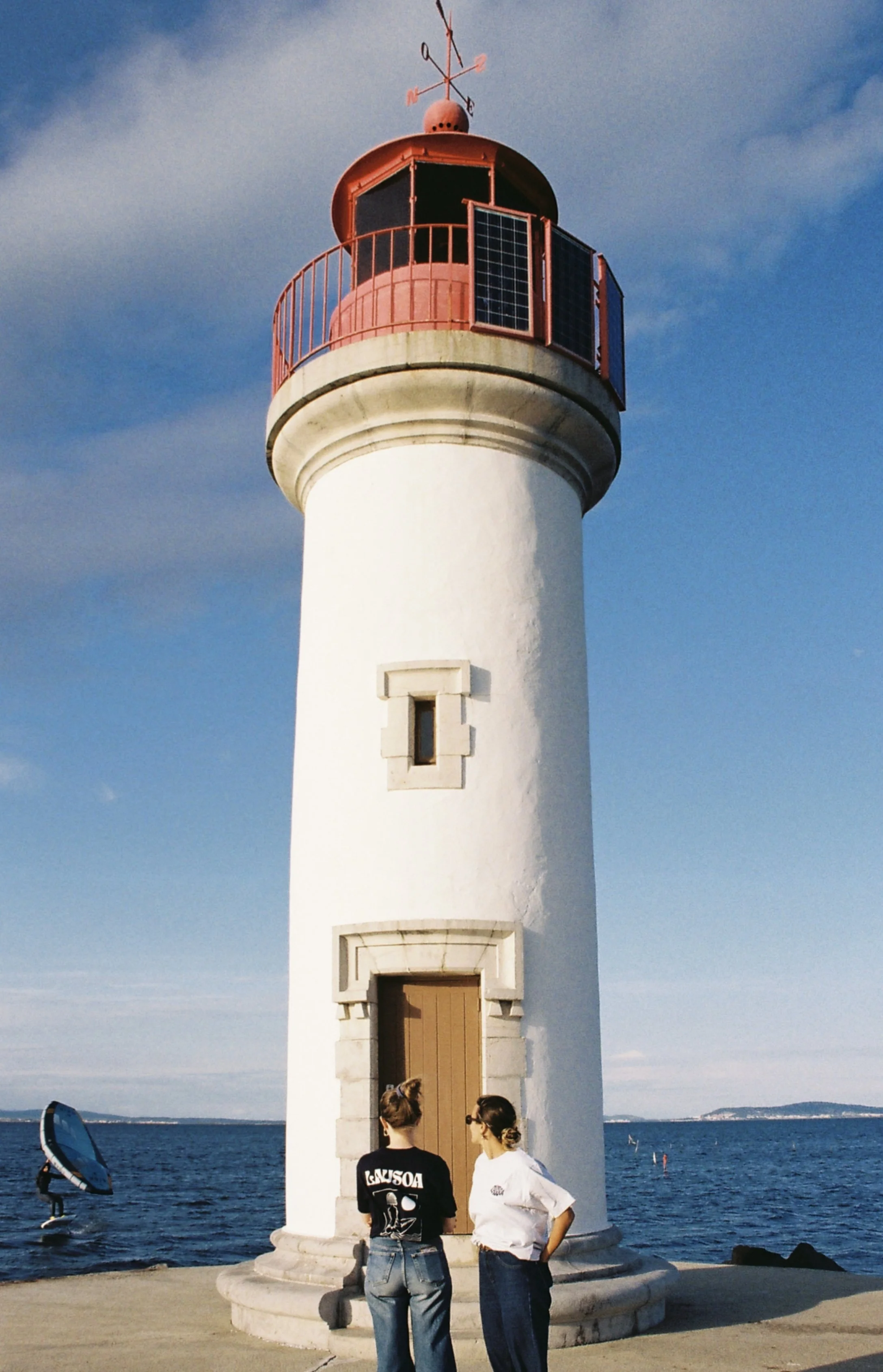Le phare blanc avec sa lanterne rouge et sa girouette, deux femmes discutent devant la mer, un stand-up paddle dans l'eau.