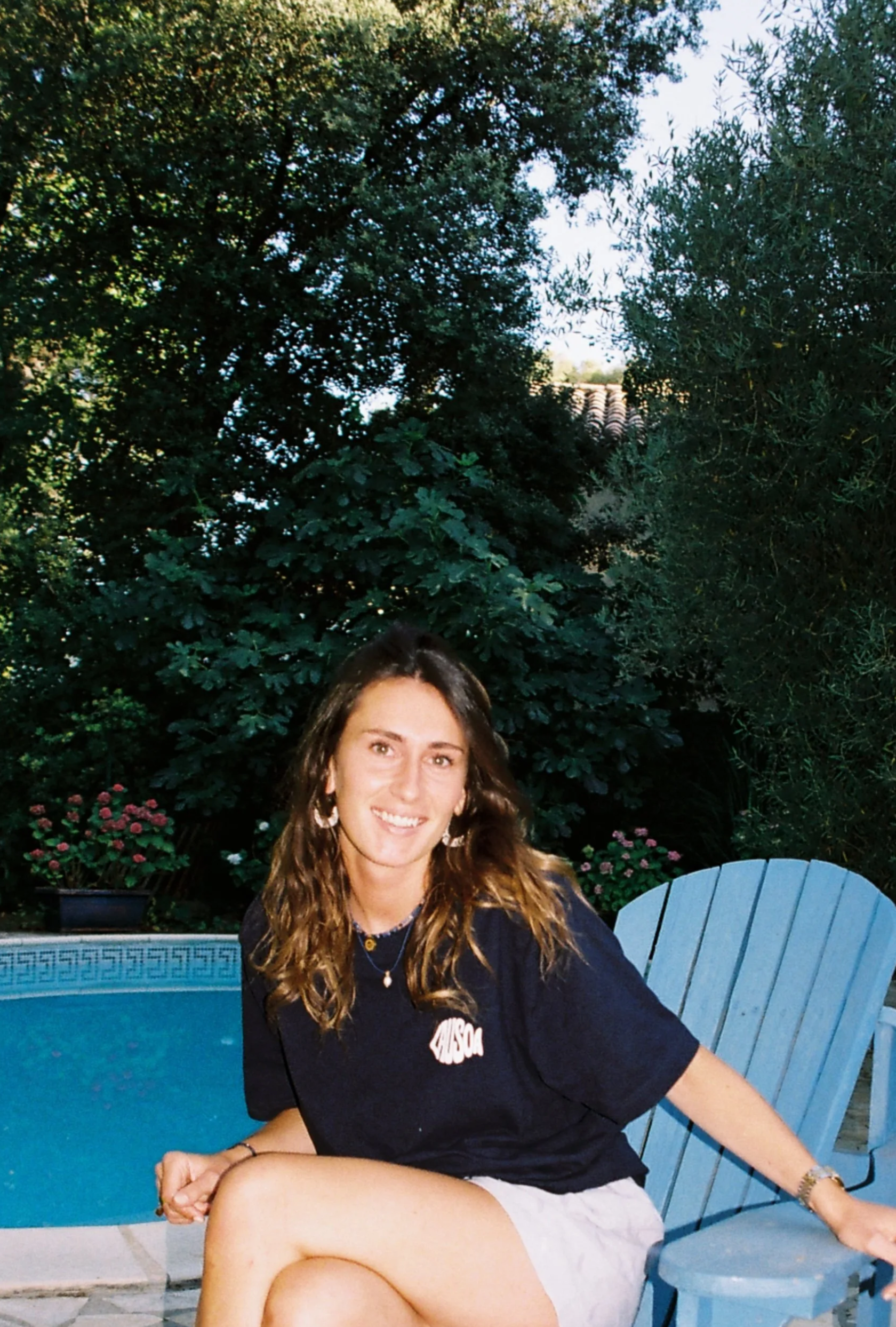 Une jeune femme assise près d'une piscine dans un jardin avec des arbres et des fleurs, souriante, portant un t-shirt bleu marine avec un logo lausoa en forme de poisson et des boucles d'oreilles.