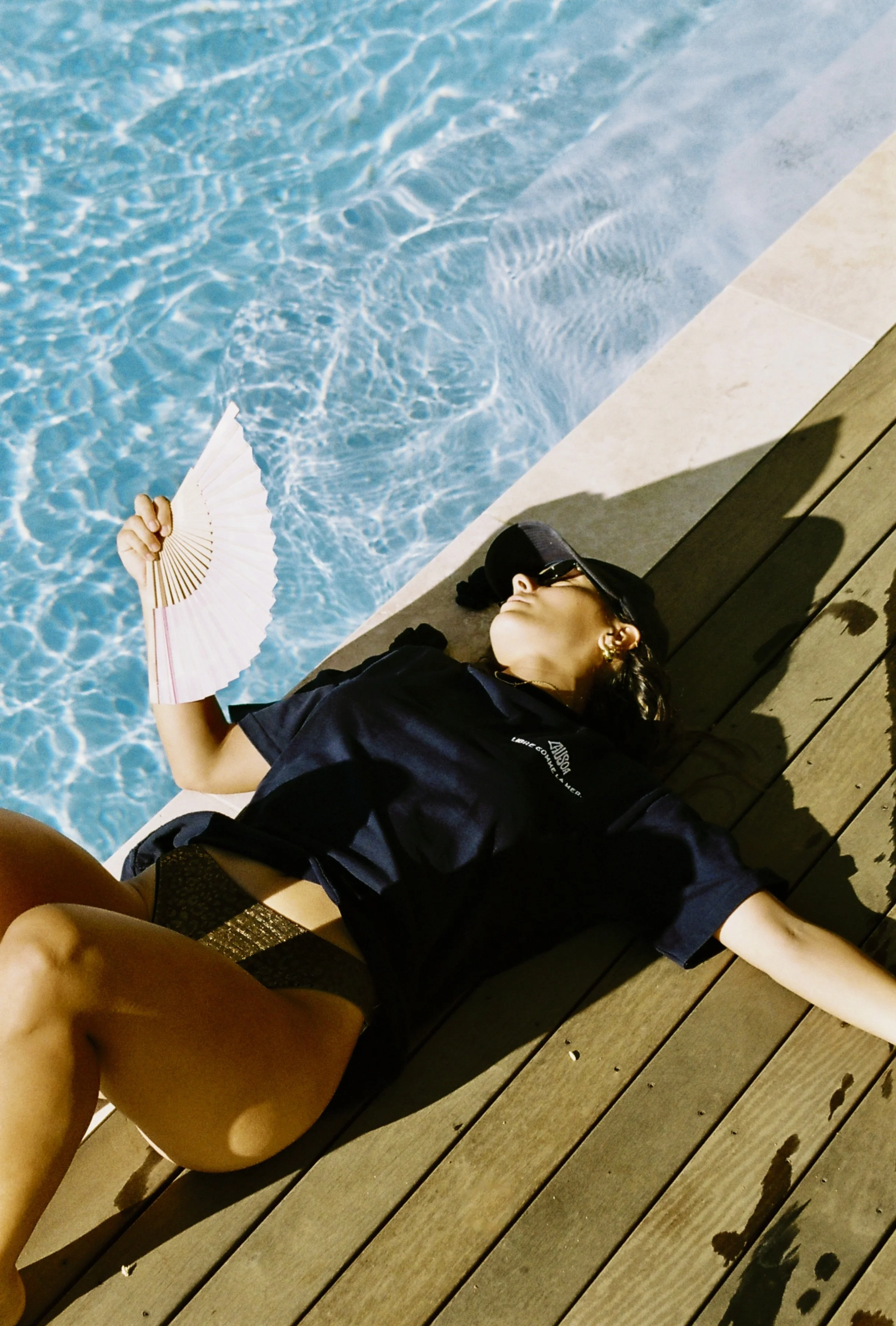 Une femme allongée sur un pont en bois à côté d'une piscine, portant une casquette noire, des lunettes de soleil, un t-shirt noir et tenant un éventail rose.