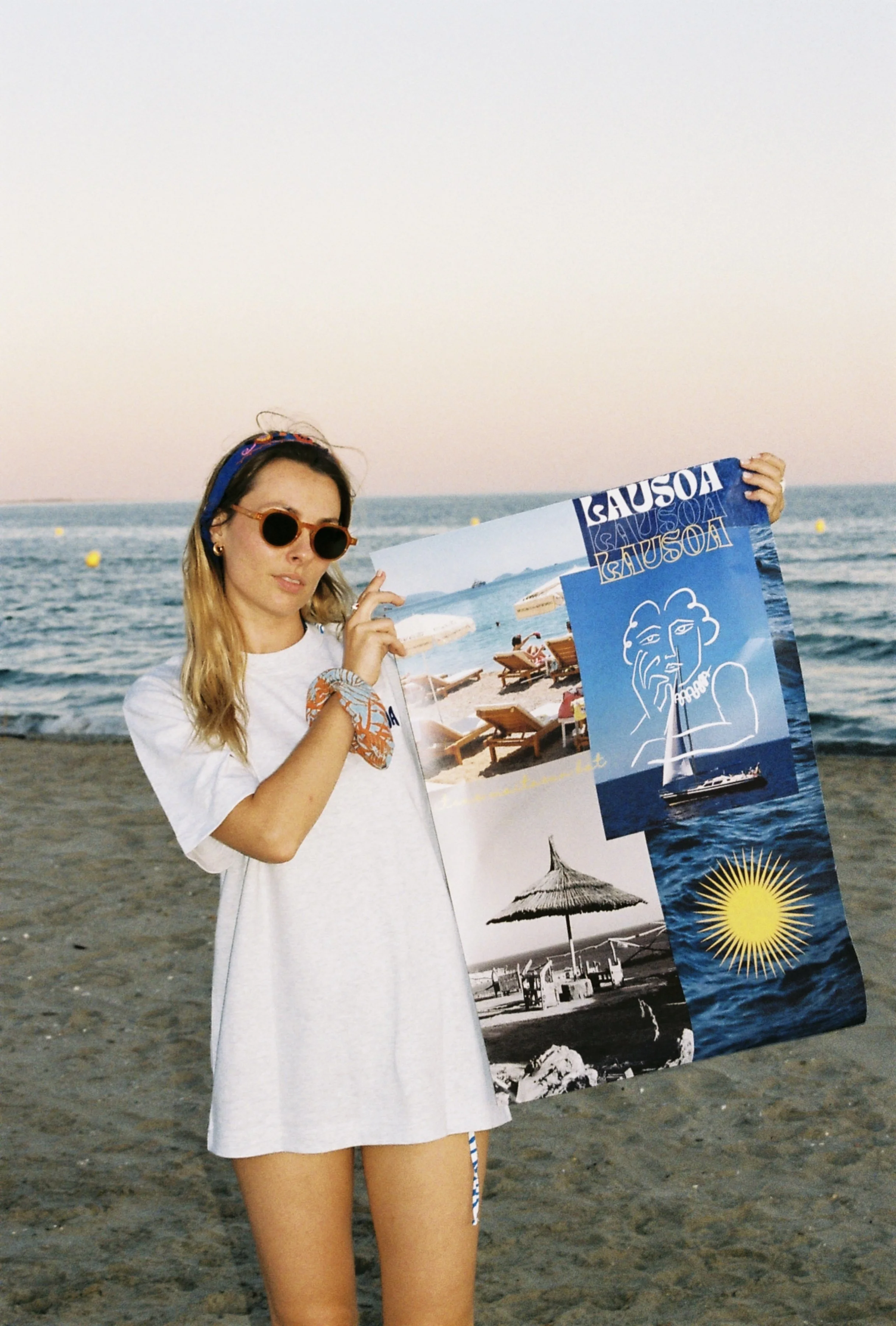 Jeune femme portant des lunettes de soleil et un bandeau, tenant une affiche publicitaire pour une station balnéaire située sur une plage avec des parasols, des chaises longues et la mer en arrière-plan.