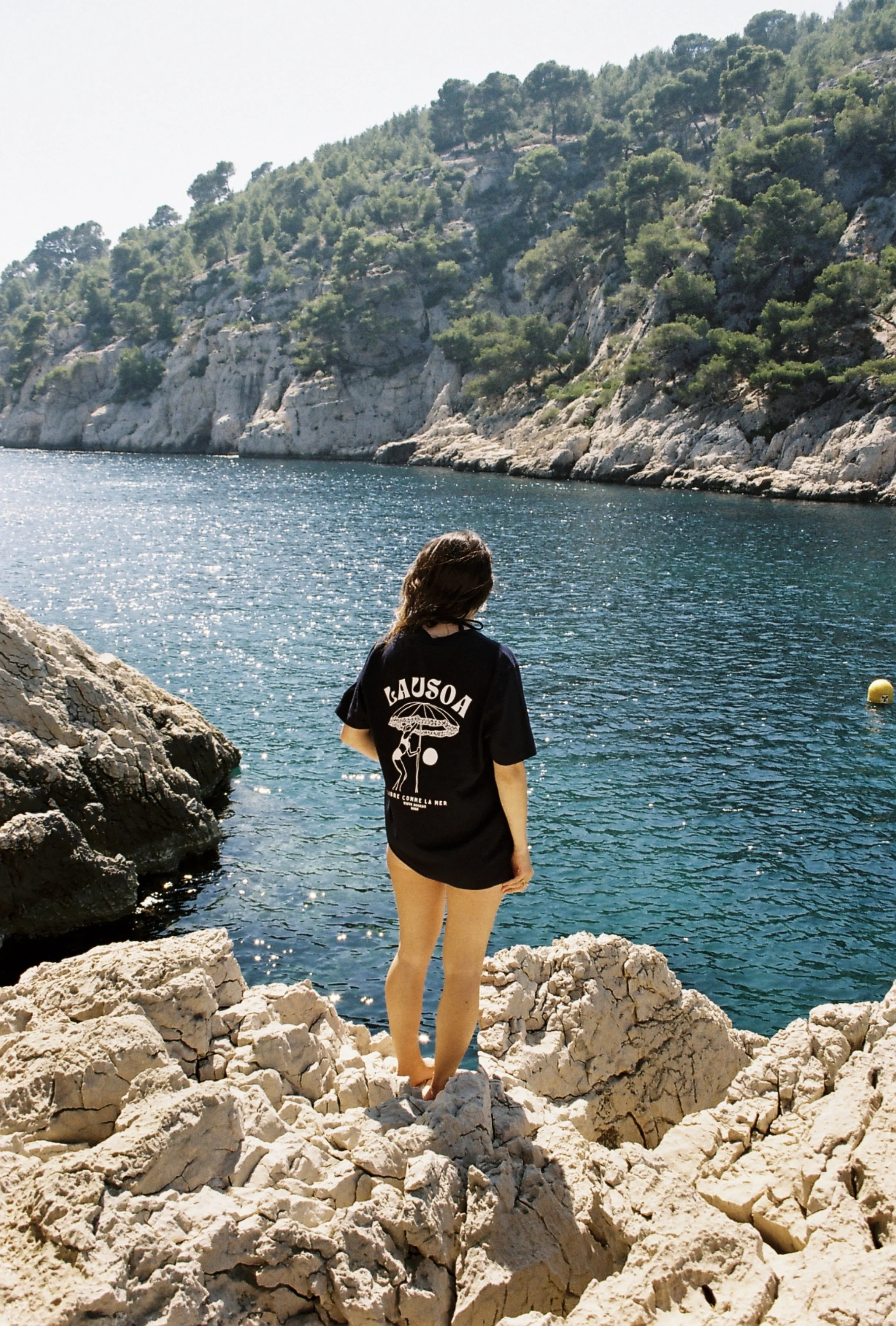 Femme avec une chemise noire qui se tient sur des rochers au bord de l'eau, avec une vue sur la mer et des collines verdoyantes en arrière-plan.