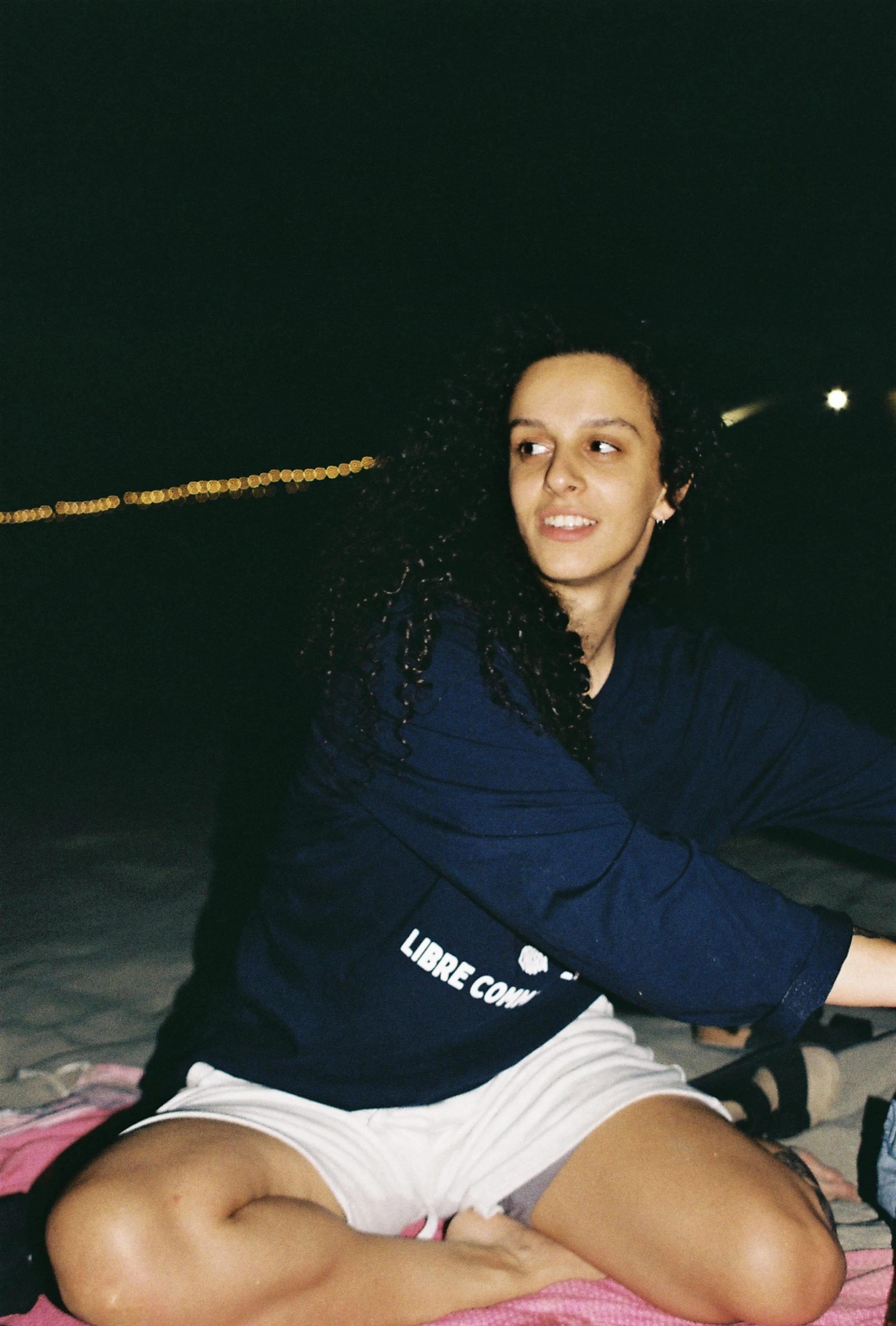 Une femme aux cheveux bouclés assise sur une serviette au bord de la mer la nuit, portant un t-shirt noir avec un texte en français et un short blanc.