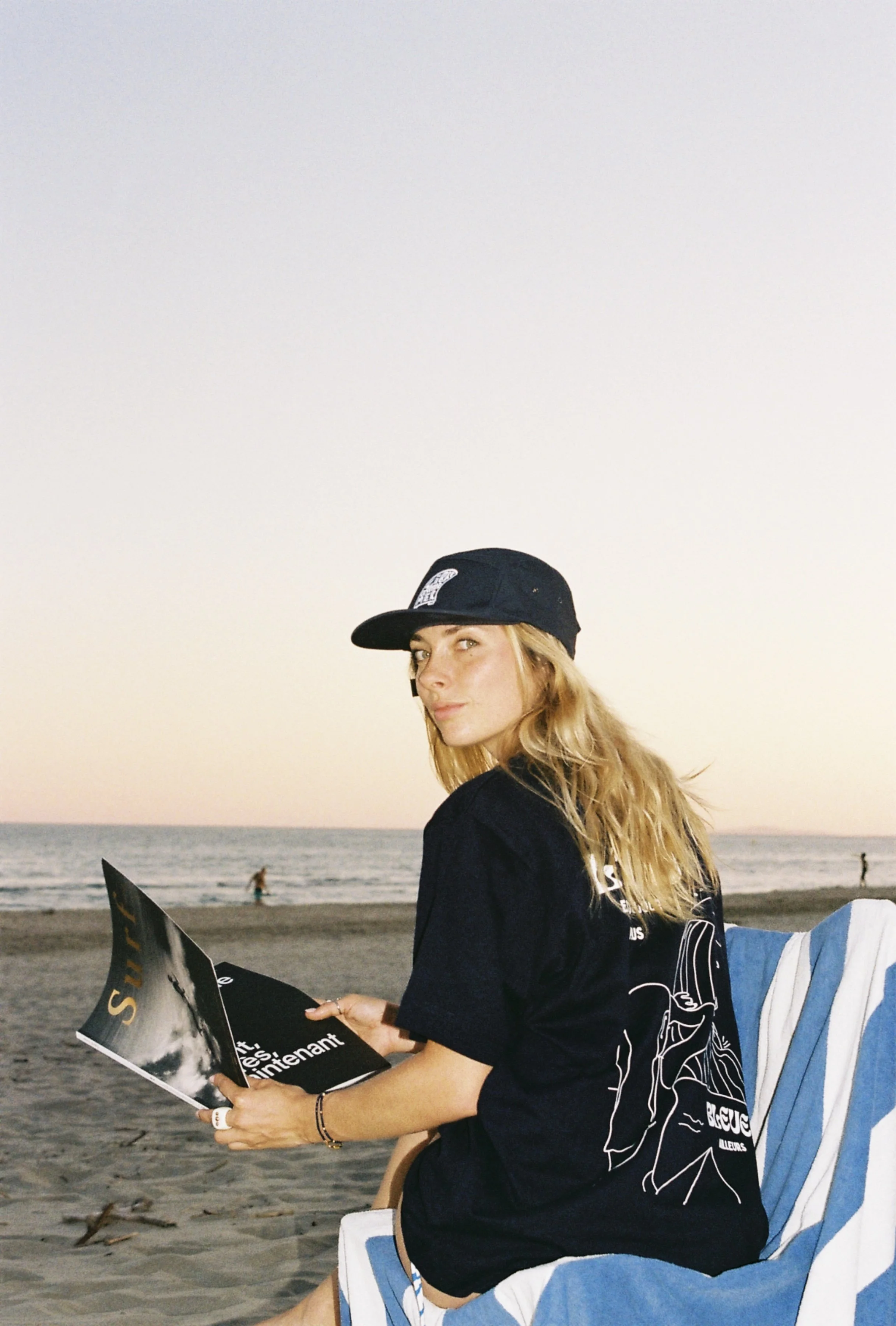 Jeune femme assise sur la plage, portant une casquette noire, un t-shirt noir, en train de lire un magazine, avec la mer en arrière-plan au coucher du soleil.
