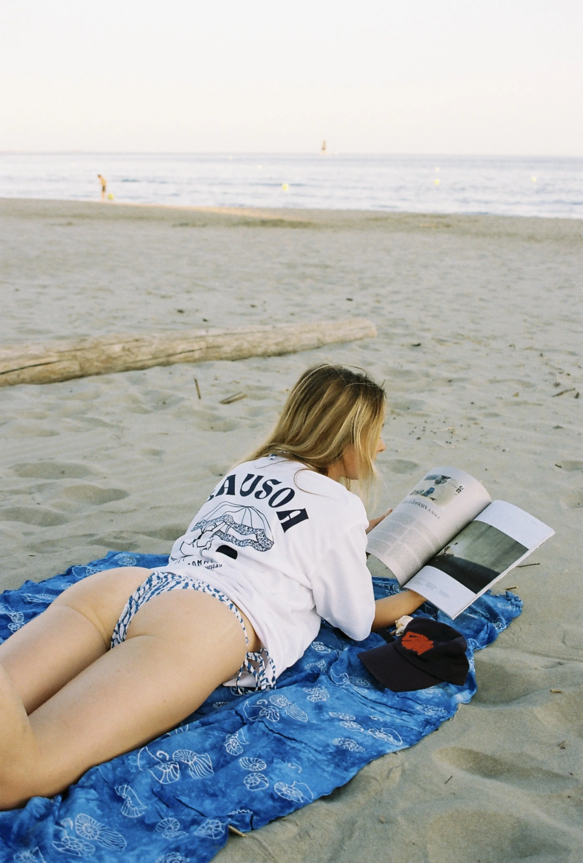 Une femme allongée sur une serviette bleue sur la plage, lire un magazine, avec la mer en arrière-plan.