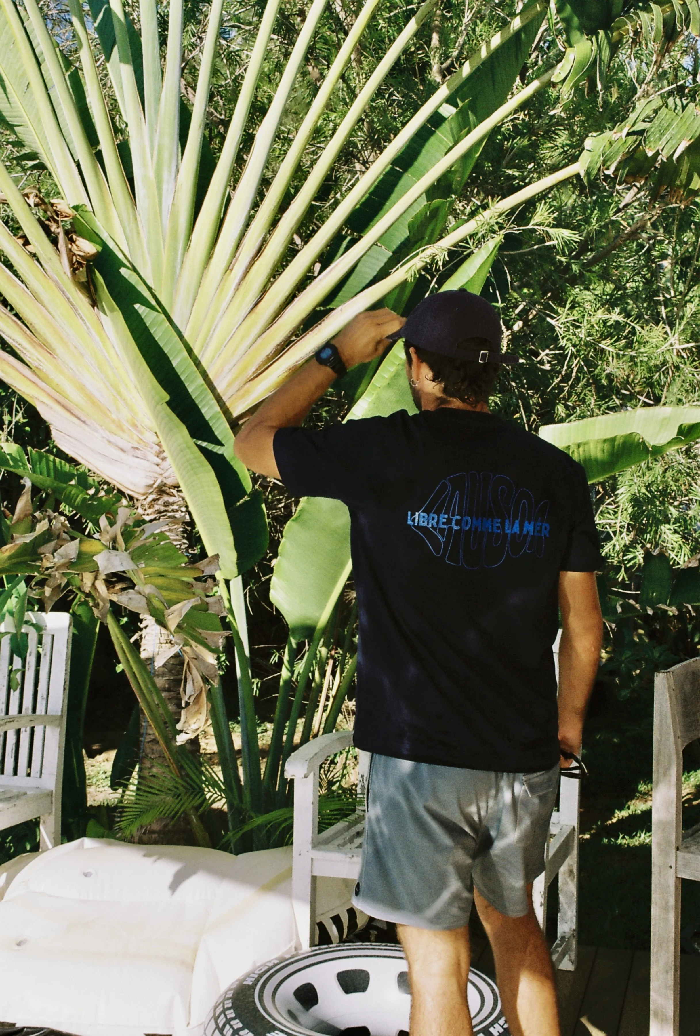 Un homme avec une casquette noire, un t-shirt noir et un short gris, vu de dos, qui touche une grande feuille de bananier dans un jardin tropical rempli de végétation verte.