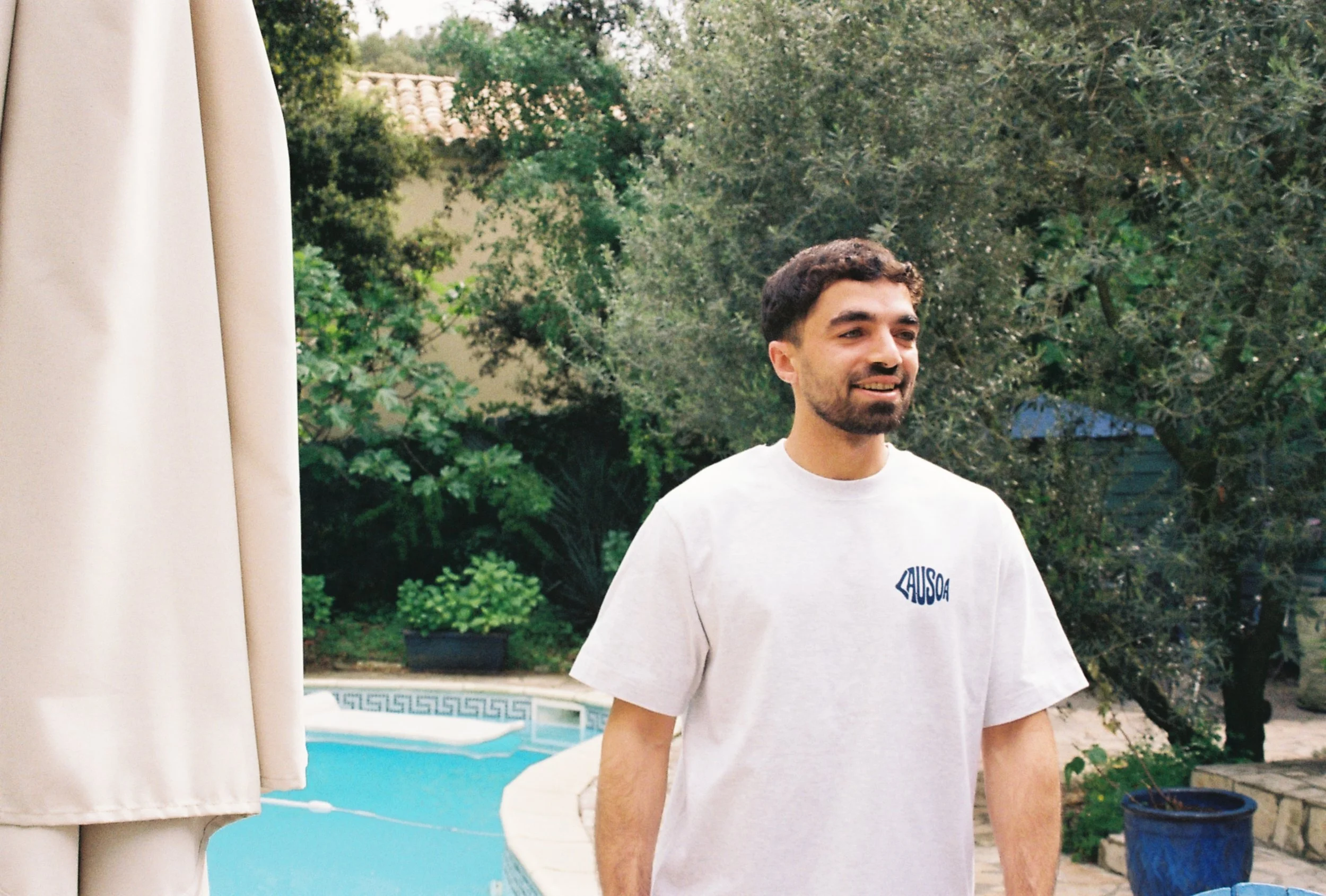 Un homme souriant se tient dans un jardin près d'une piscine, portant un t-shirt blanc avec un logo bleu.