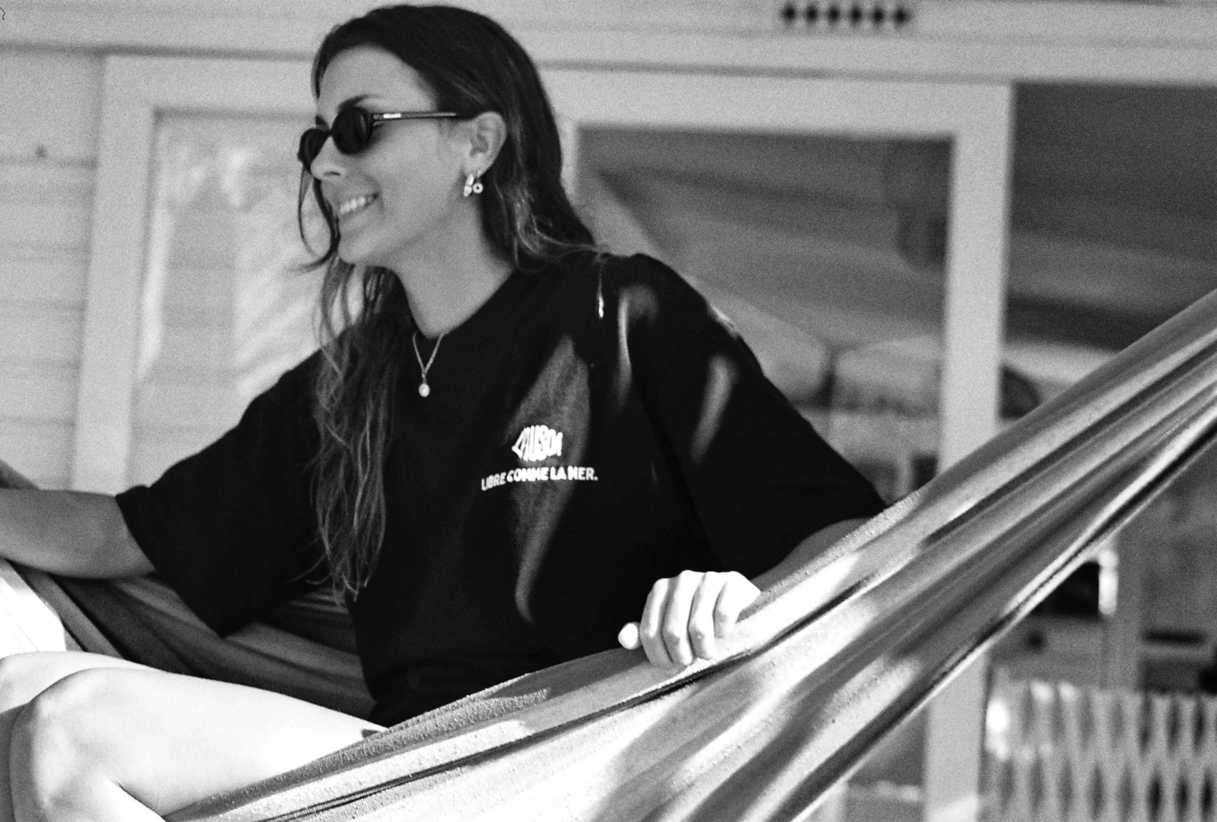 Une femme souriante portant des lunettes de soleil noires, des boucles d'oreilles et un collier, assise sur une voile de bateau en bois, avec un environnement intérieur en bois visible en arrière-plan.