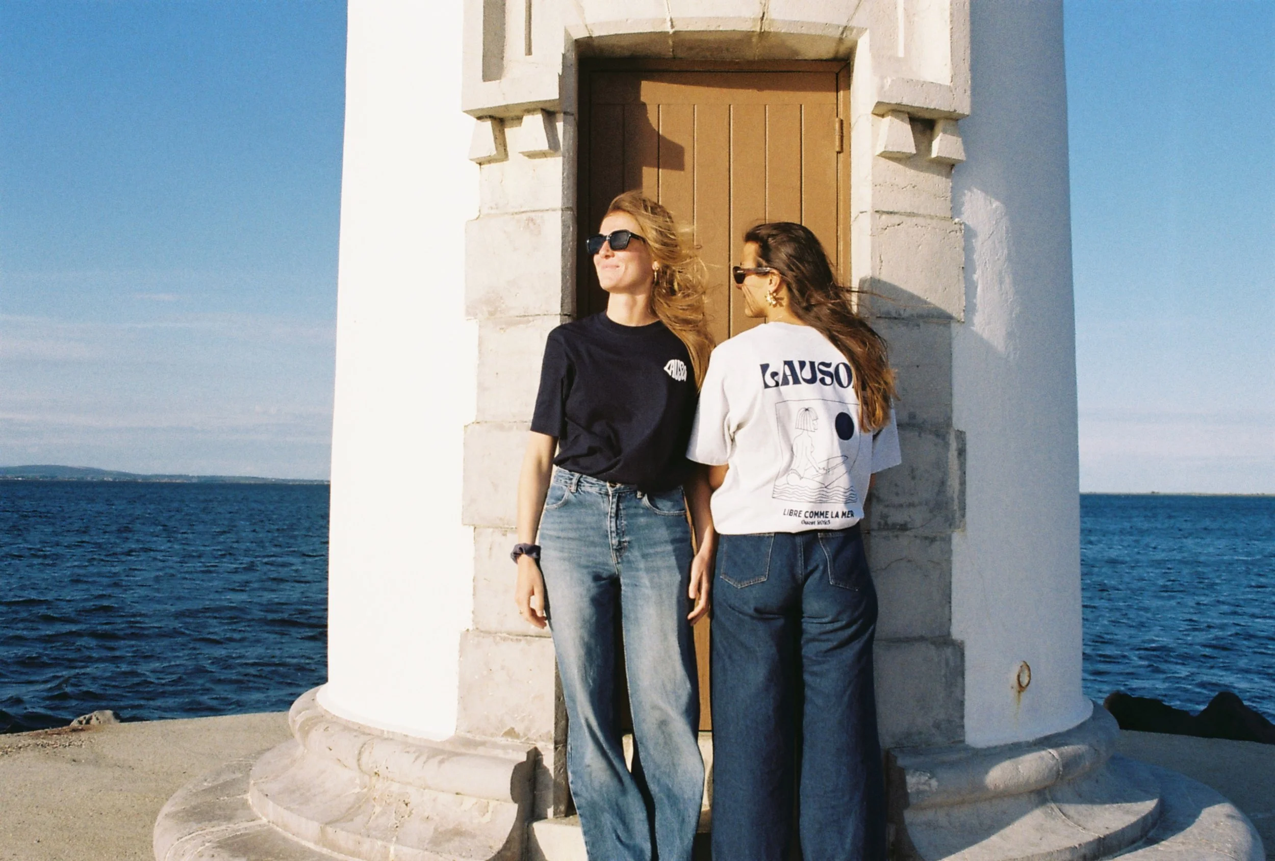 Deux femmes debout devant une petite structure en pierre, près de la mer, portant des t-shirts décontractés et des jeans, avec un ciel bleu clair en arrière-plan.
