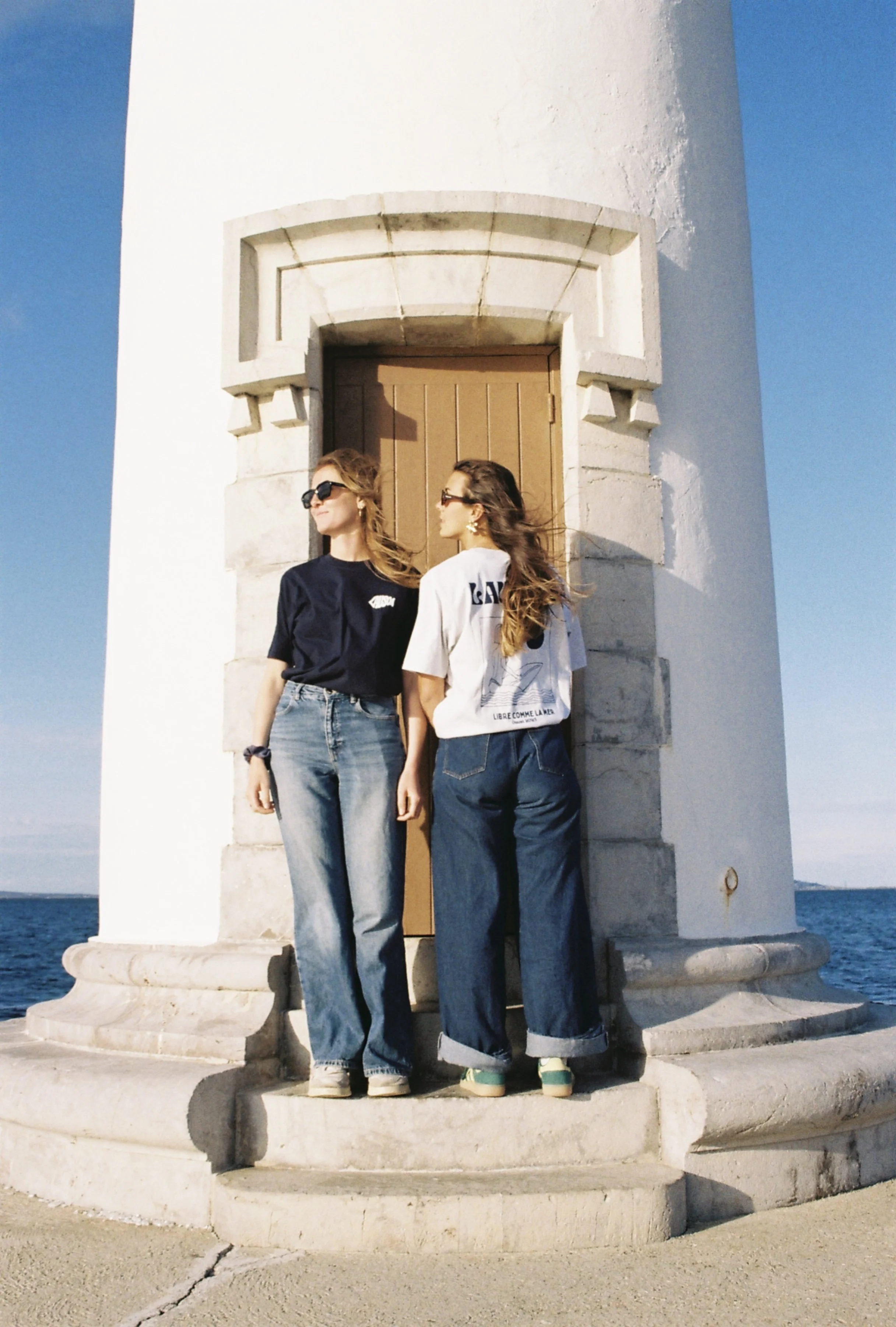 Deux femmes portant des jeans et des t-shirts décontractés se tenant devant une petite tour blanche en bord de mer, sous un ciel bleu clair.
