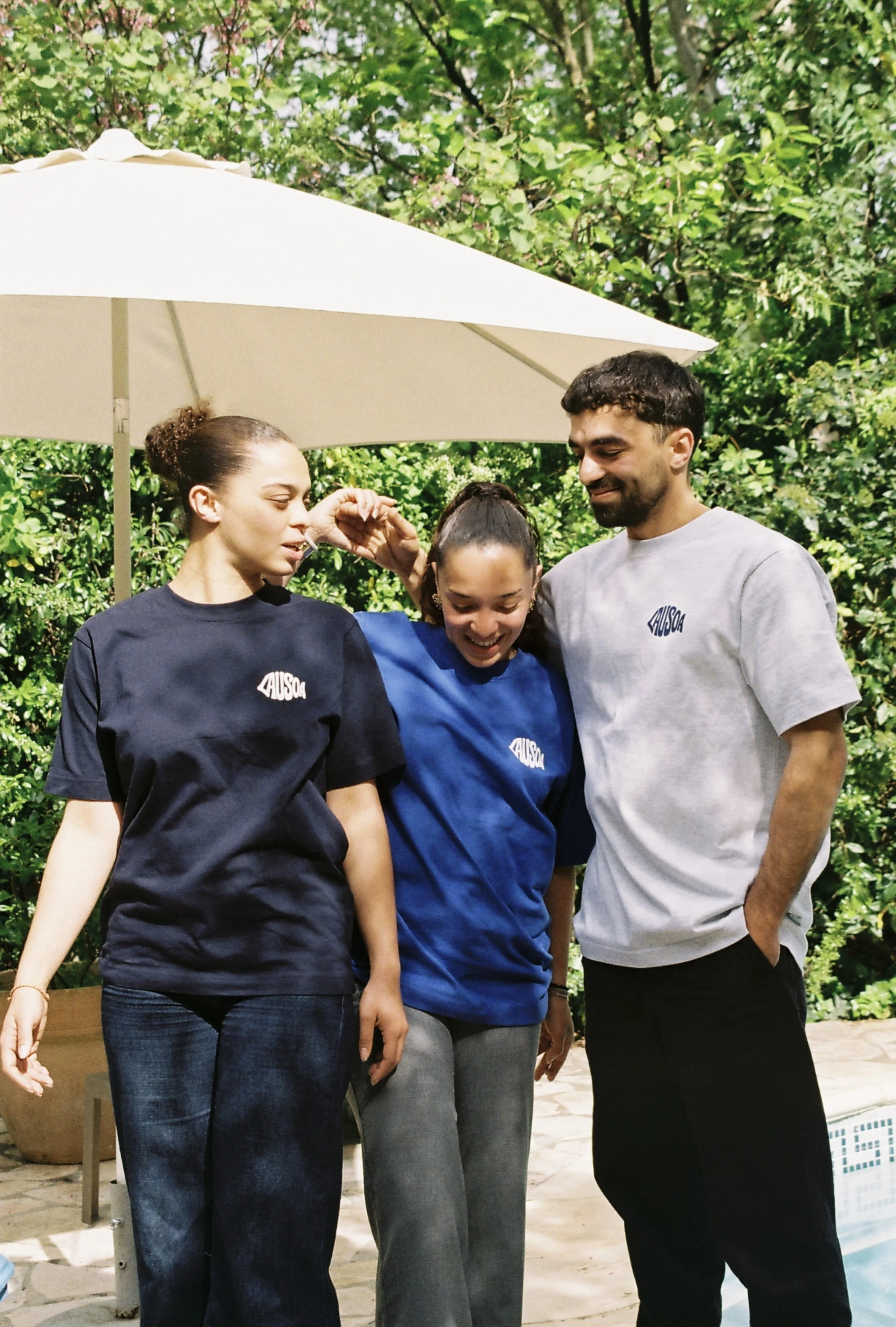 Trois personnes portant des t-shirts avec le logo 'AUSOA' : deux femmes et un homme, souriants et en interaction, à l'extérieur sous un parasol, entourés de verdure.