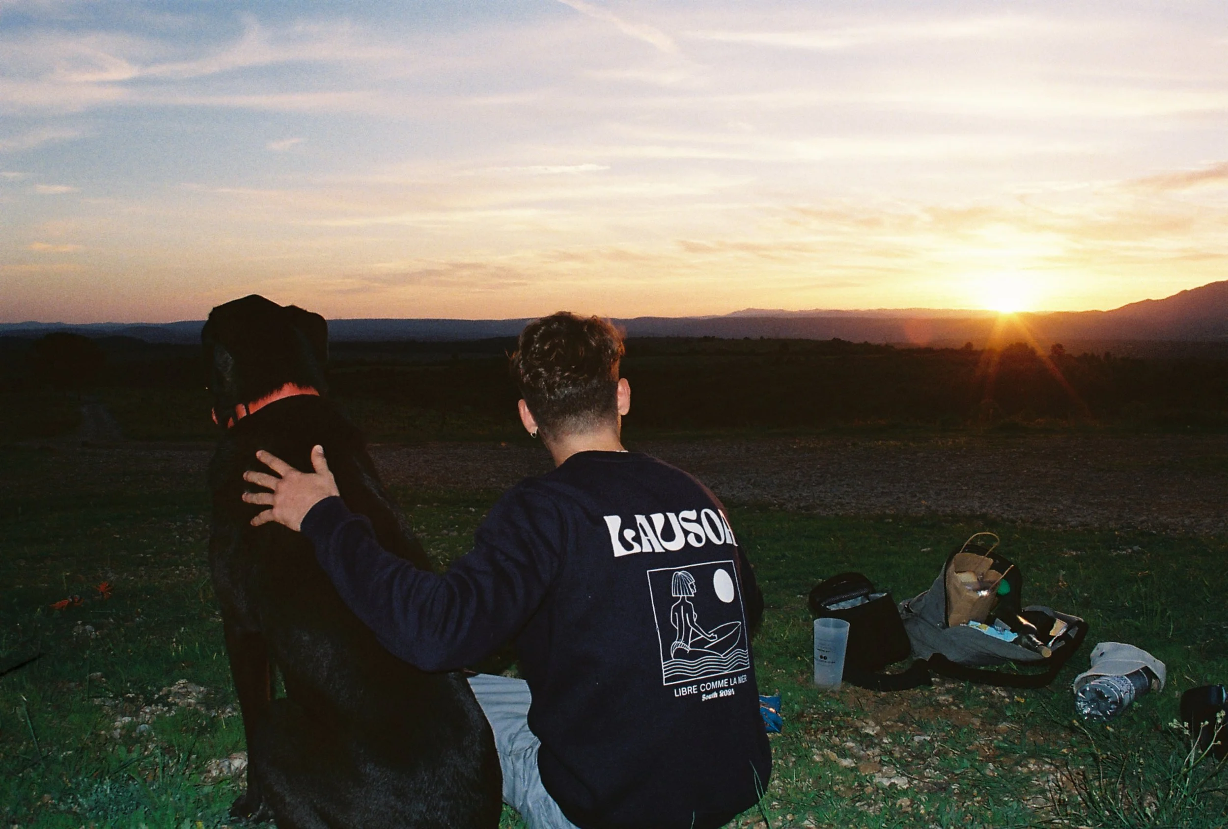 Une personne avec un tatouage sur le dos, portant une veste sombre, assise sur l'herbe, caressant un chien noir, lors d'un coucher de soleil dans un paysage rural.