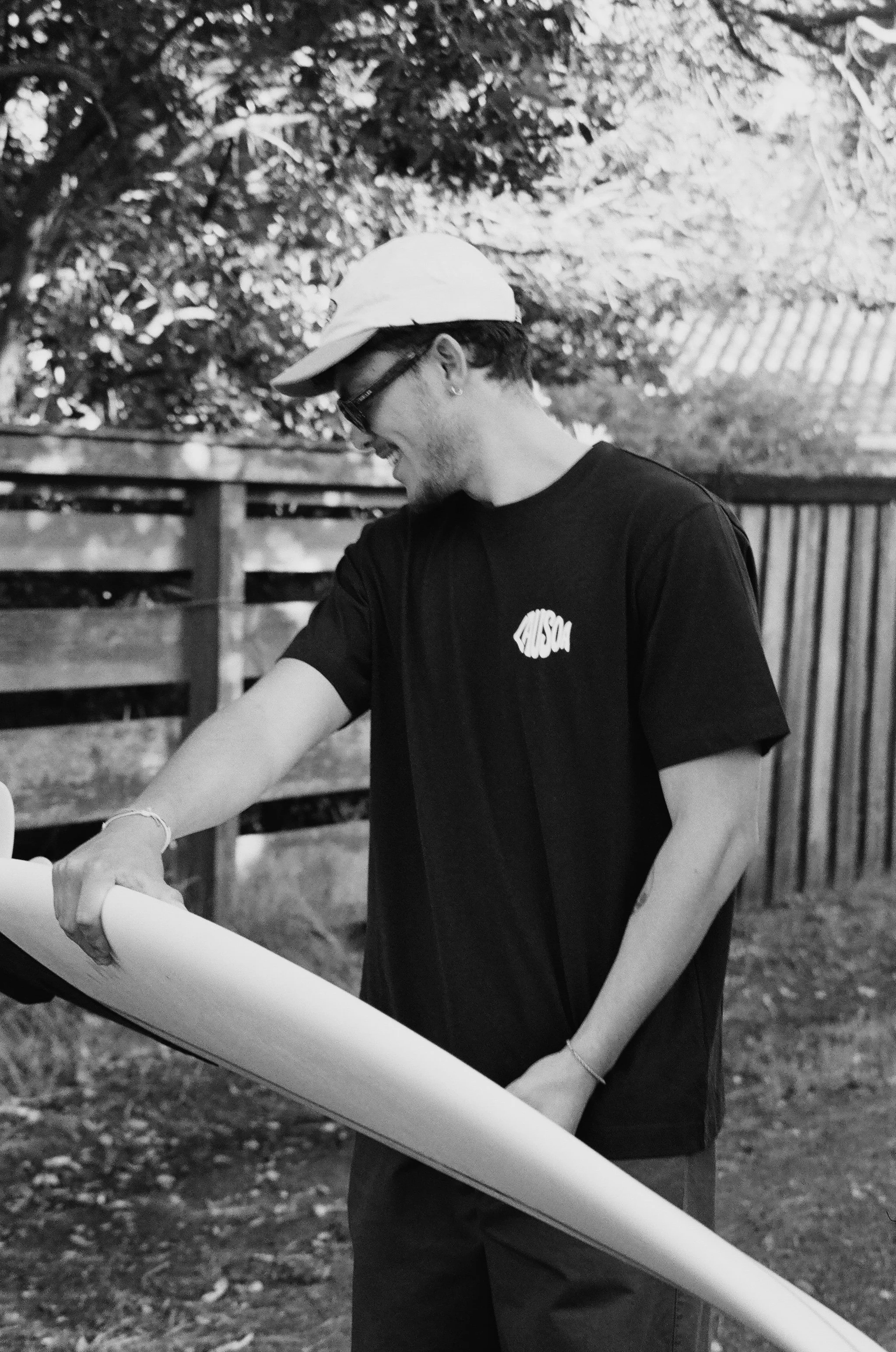 Jeune homme portant un t-shirt noir avec l'inscription «AUSMA», une casquette blanche et des lunettes de soleil, souriant tout en tenant un plan ou un papier, dans un jardin en plein air.