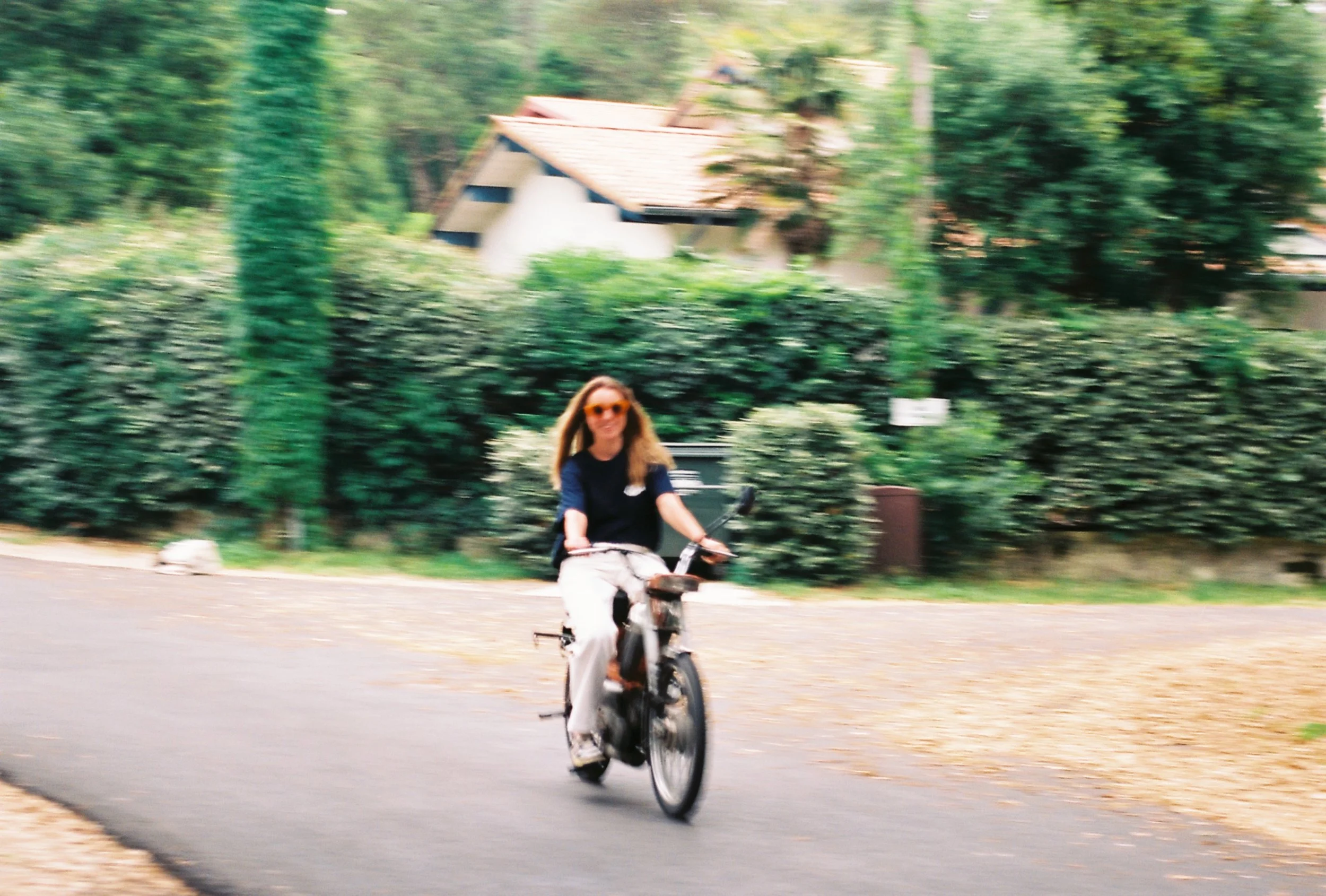 Femme souriante portant des lunettes de soleil, faisant du vélo sur une voie pavée dans un environnement verdoyant avec des maisons en arrière-plan.