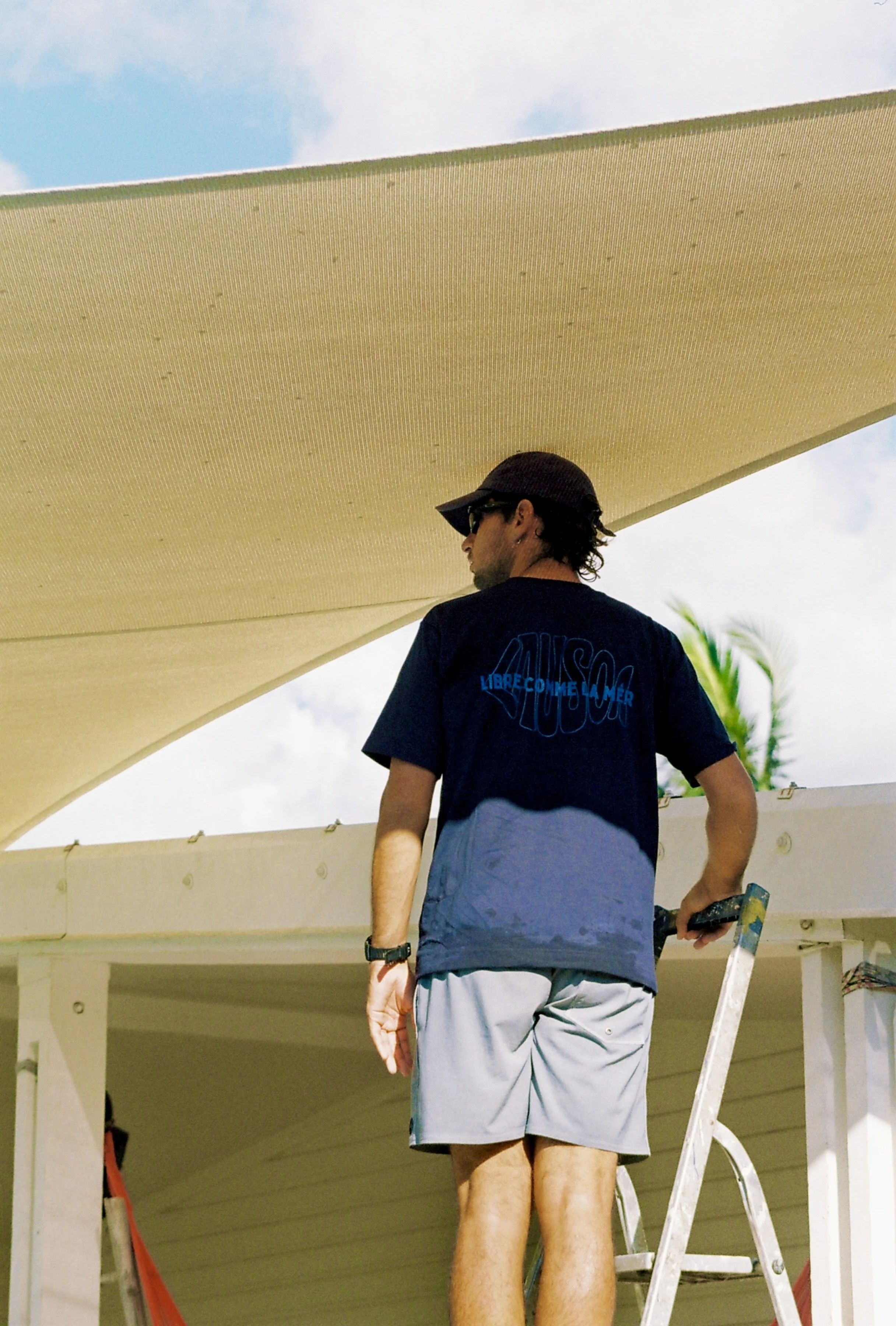 Un homme debout sur une échelle, vu de dos, portant un t-shirt noir avec un texte en français, shorts gris, sous un plafond en béton près de la mer, avec un palmier en arrière-plan.