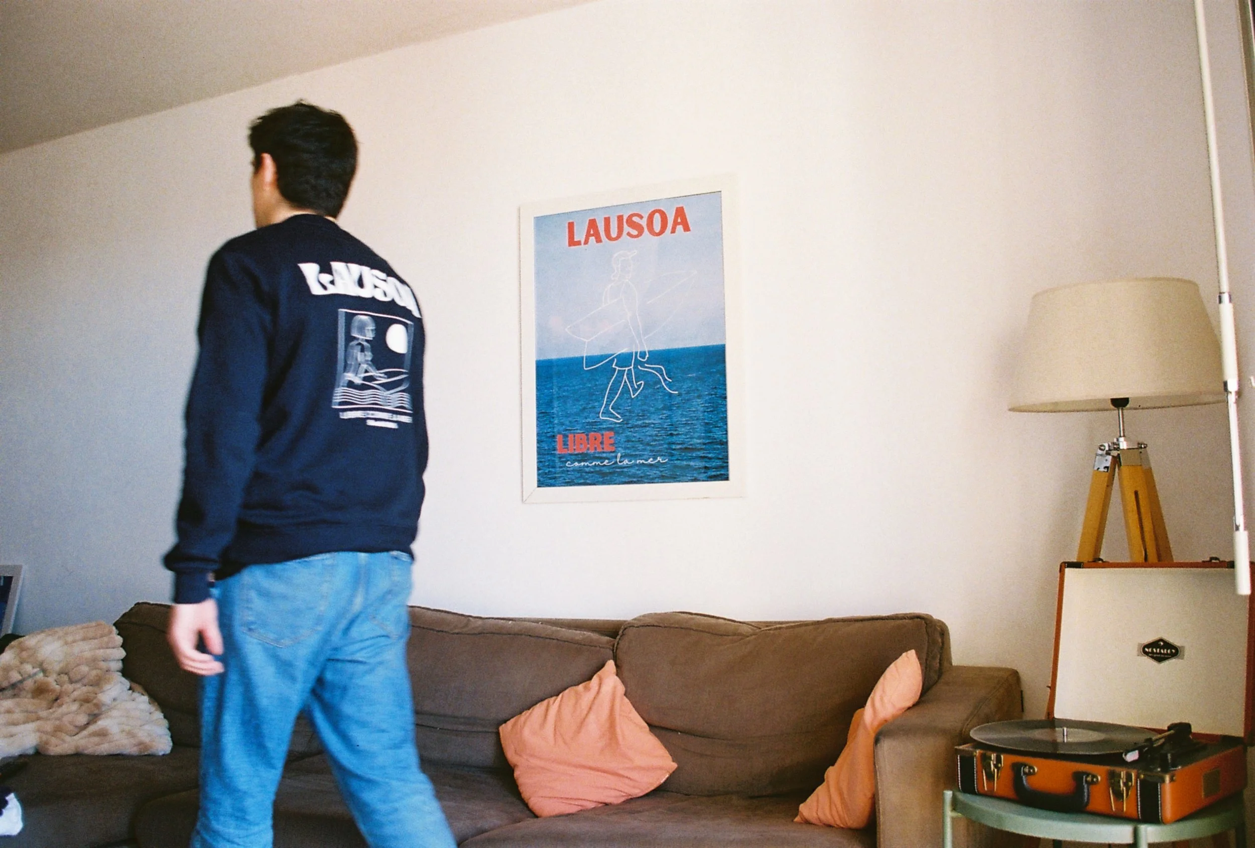 Un homme avec un blouson crème et un pantalon bleu clair dans un salon, mur avec une affiche de Lausoa et un canapé marron avec des coussins rose et beige, lampe avec abajour beige et tourne-disque orange.
