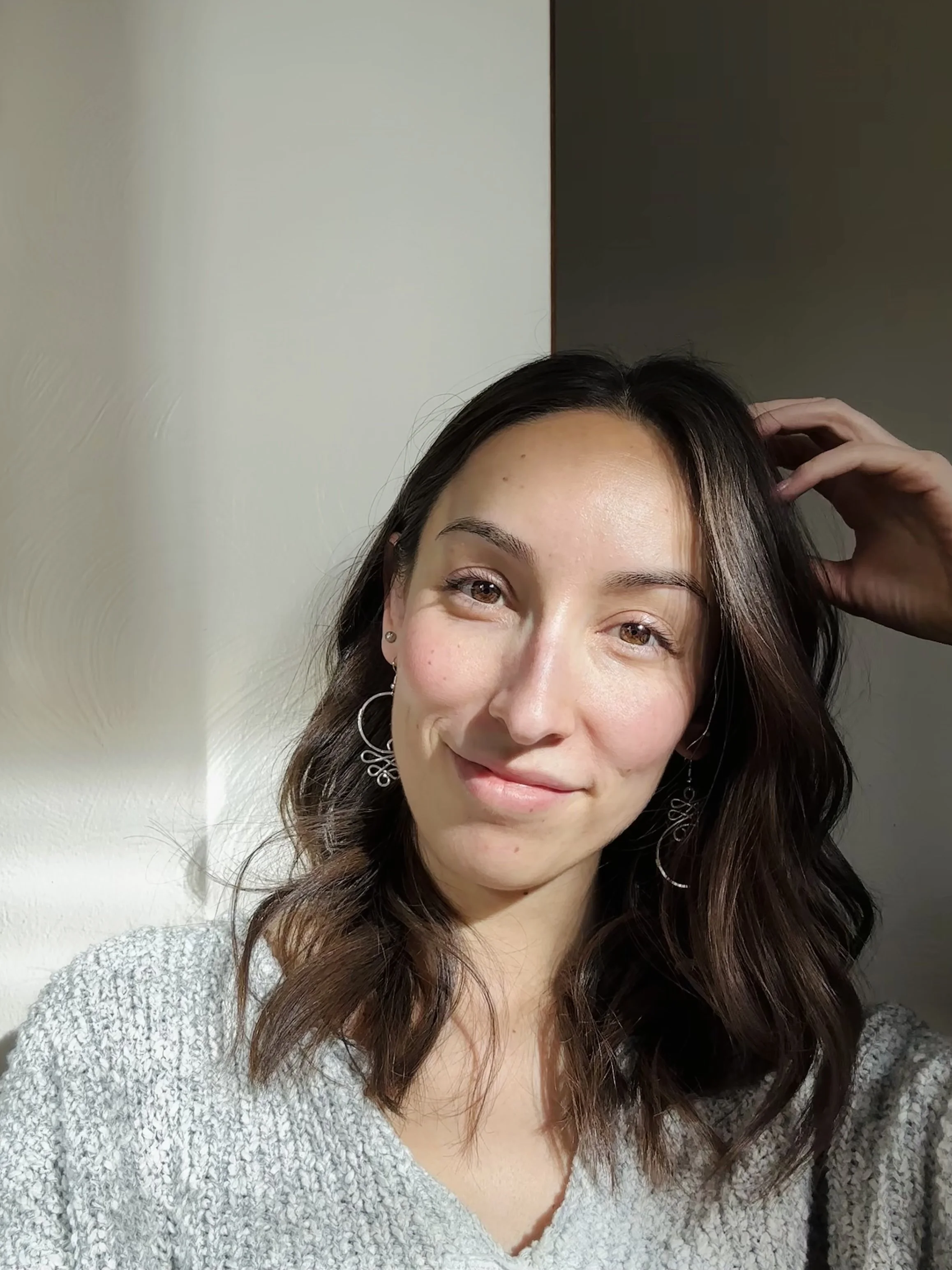 A woman with dark wavy hair, wearing earrings and a light gray sweater, smiling at the camera with sunlight illuminating her face.