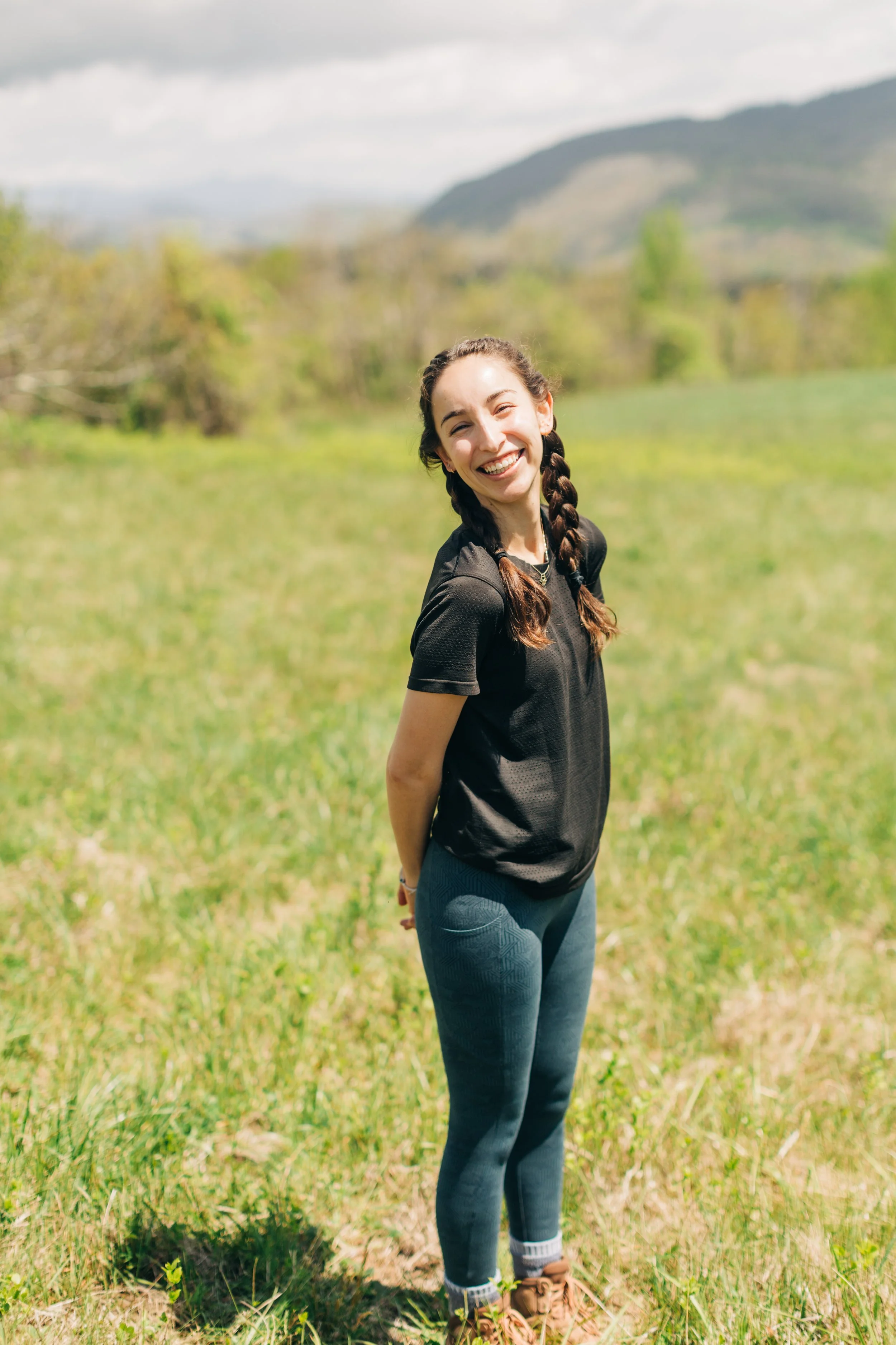 A young woman with braided hair, wearing a black athletic shirt, blue leggings, and brown shoes, standing in a grassy field with a mountain in the background, smiling at the camera.