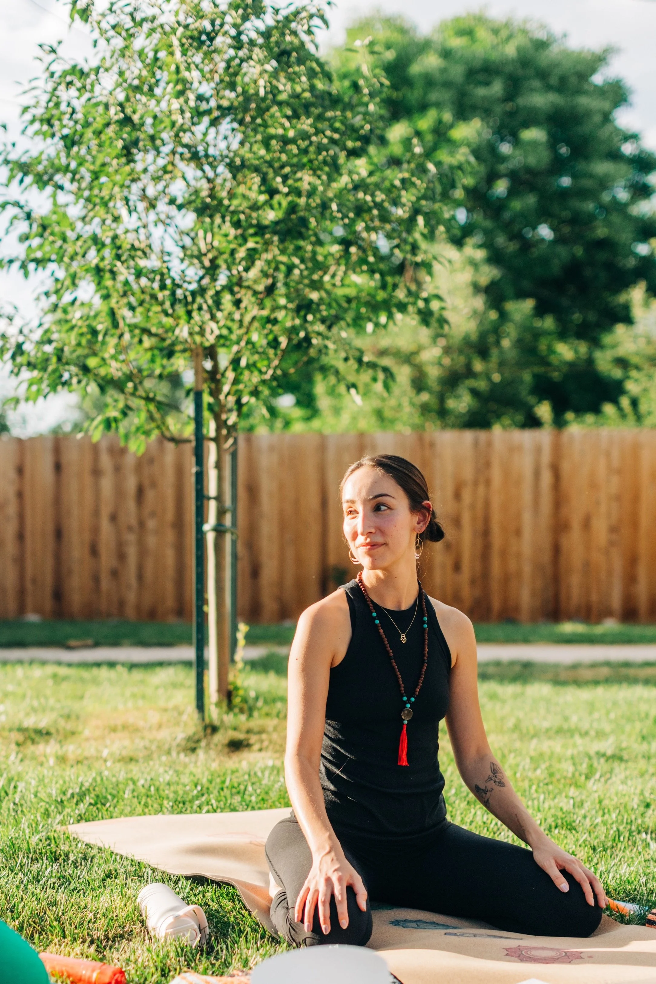 A woman practicing yoga outdoors on a mat in a backyard with green grass, a tree, and a wooden fence.