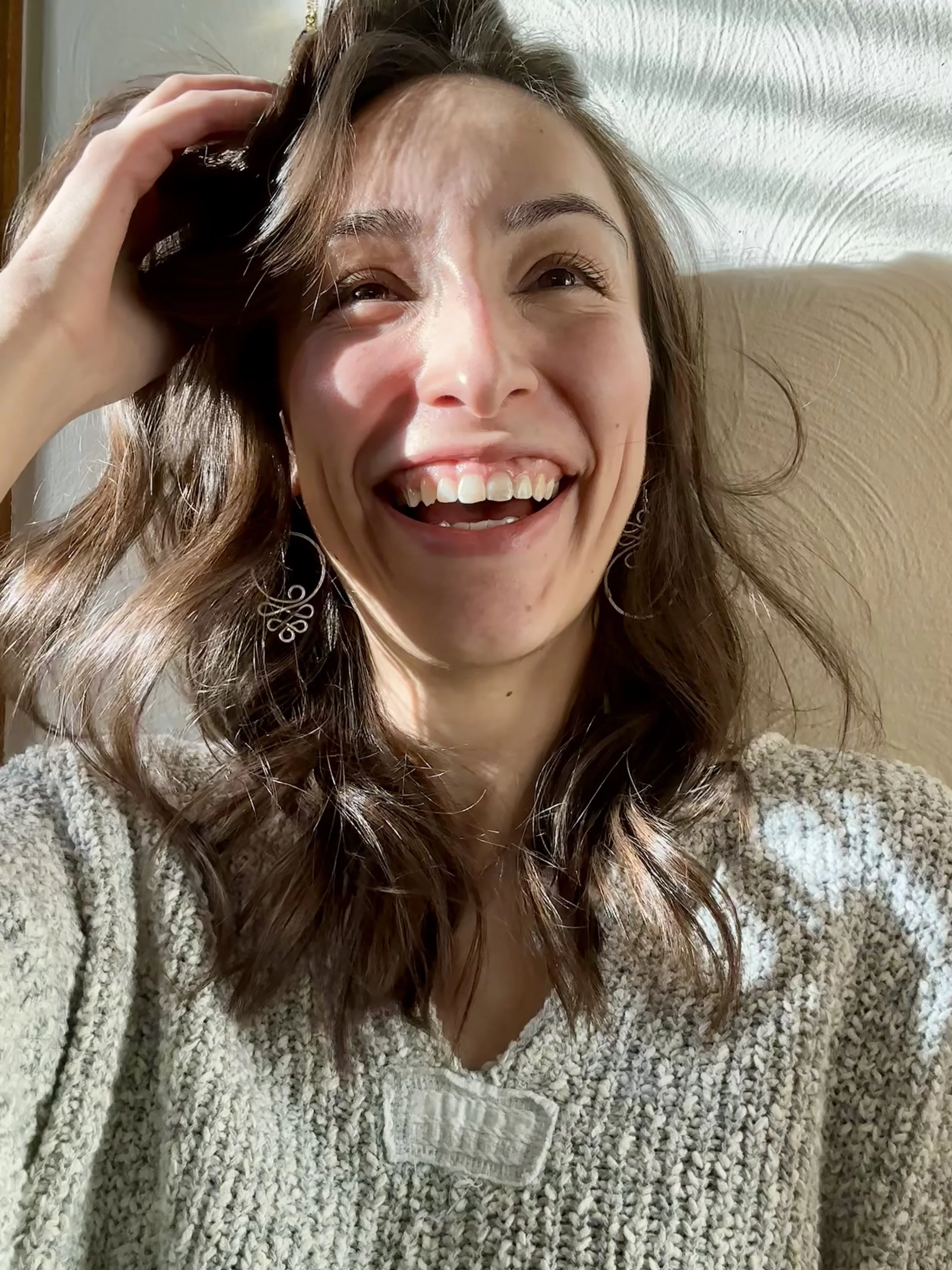 A woman with wavy brown hair smiling brightly, touching her hair, wearing a cozy beige sweater with a textured pattern and large earrings, sitting against a light-colored backdrop.