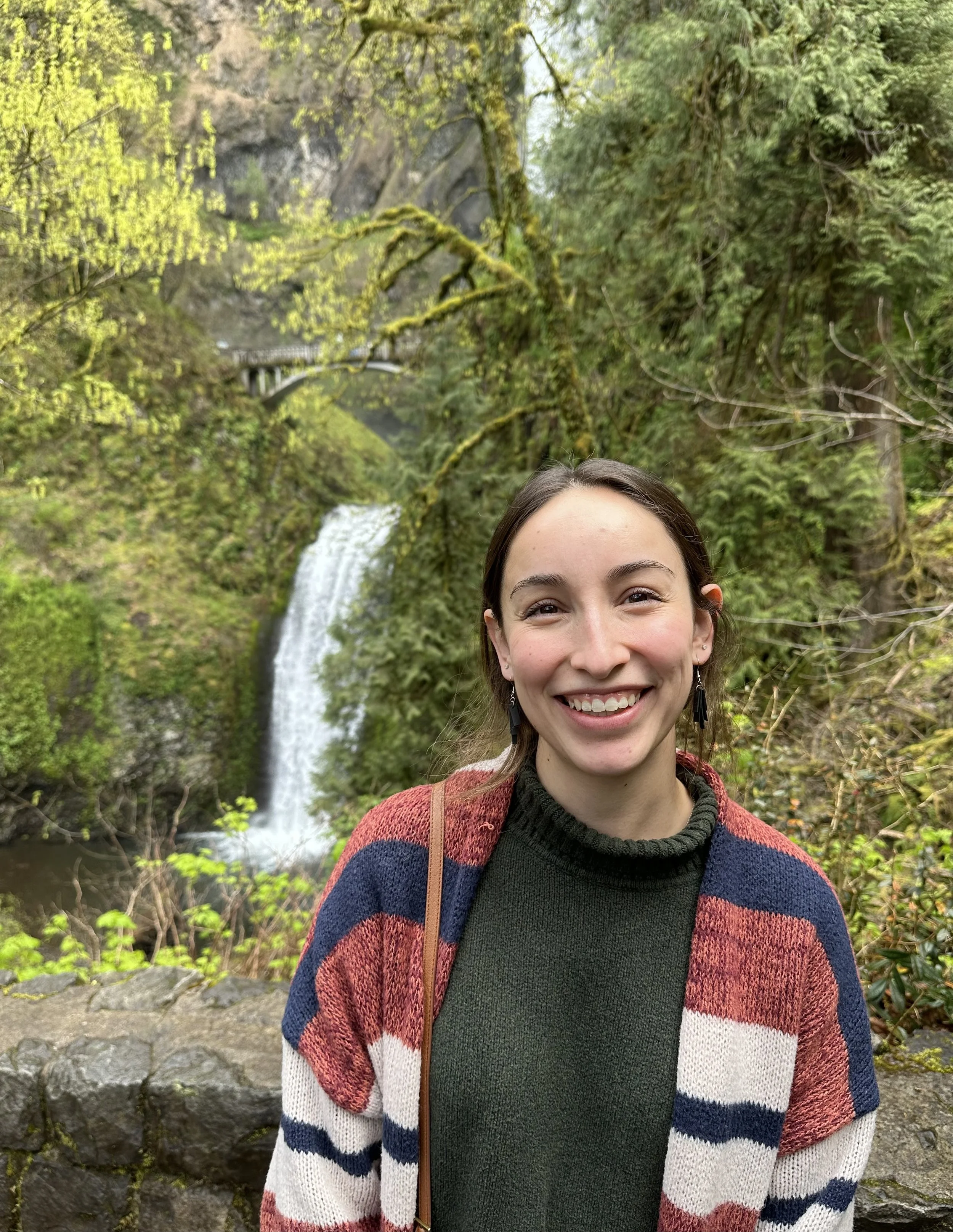 Smiling woman standing in front of a waterfall and lush green trees in a forest.
