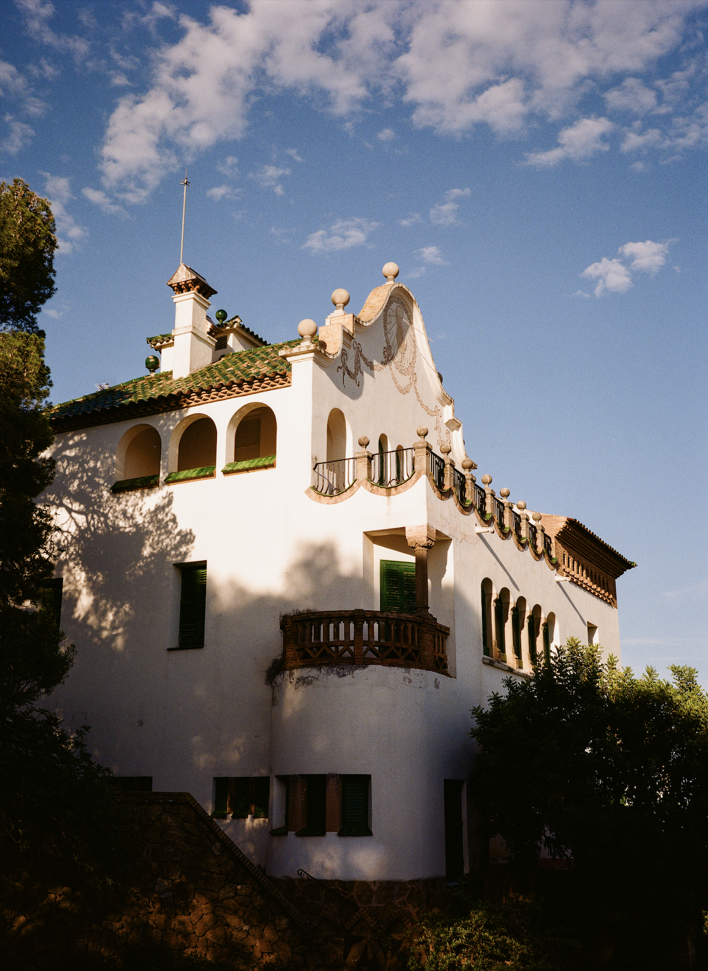 A Spanish-style white building with arched windows, decorative balconies, and a terracotta roof, set against a blue sky with scattered clouds.