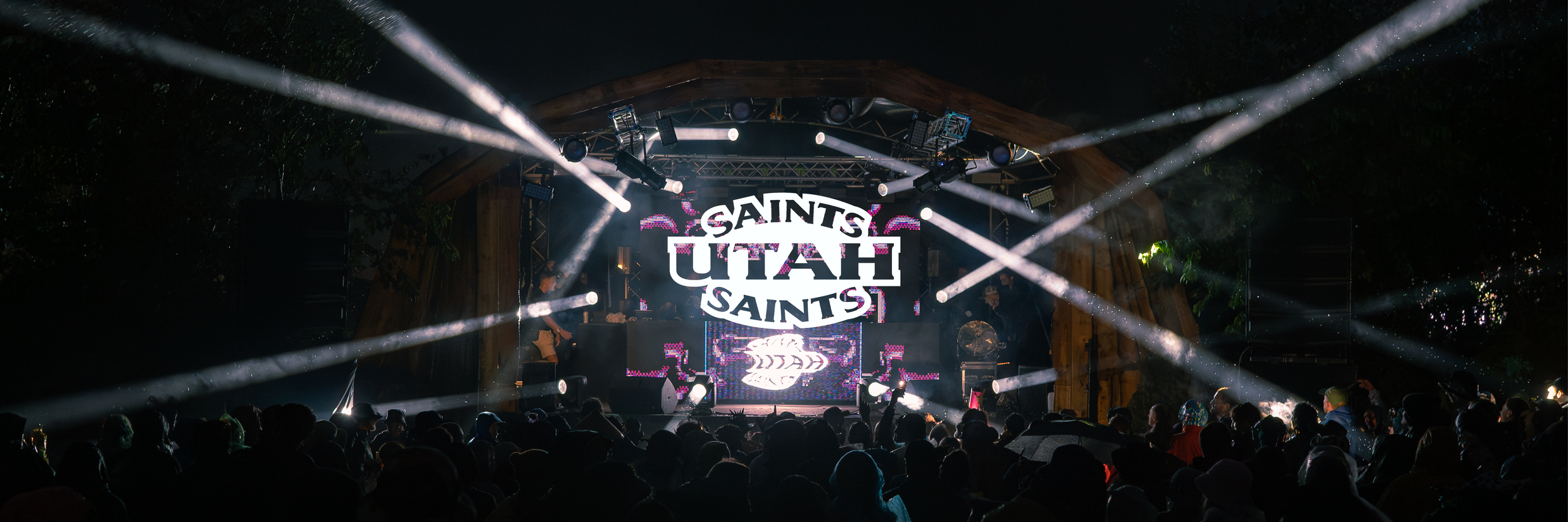 Nighttime outdoor concert with a stage, bright lights, and a crowd, featuring a large screen displaying the words 'Saints Utah'.