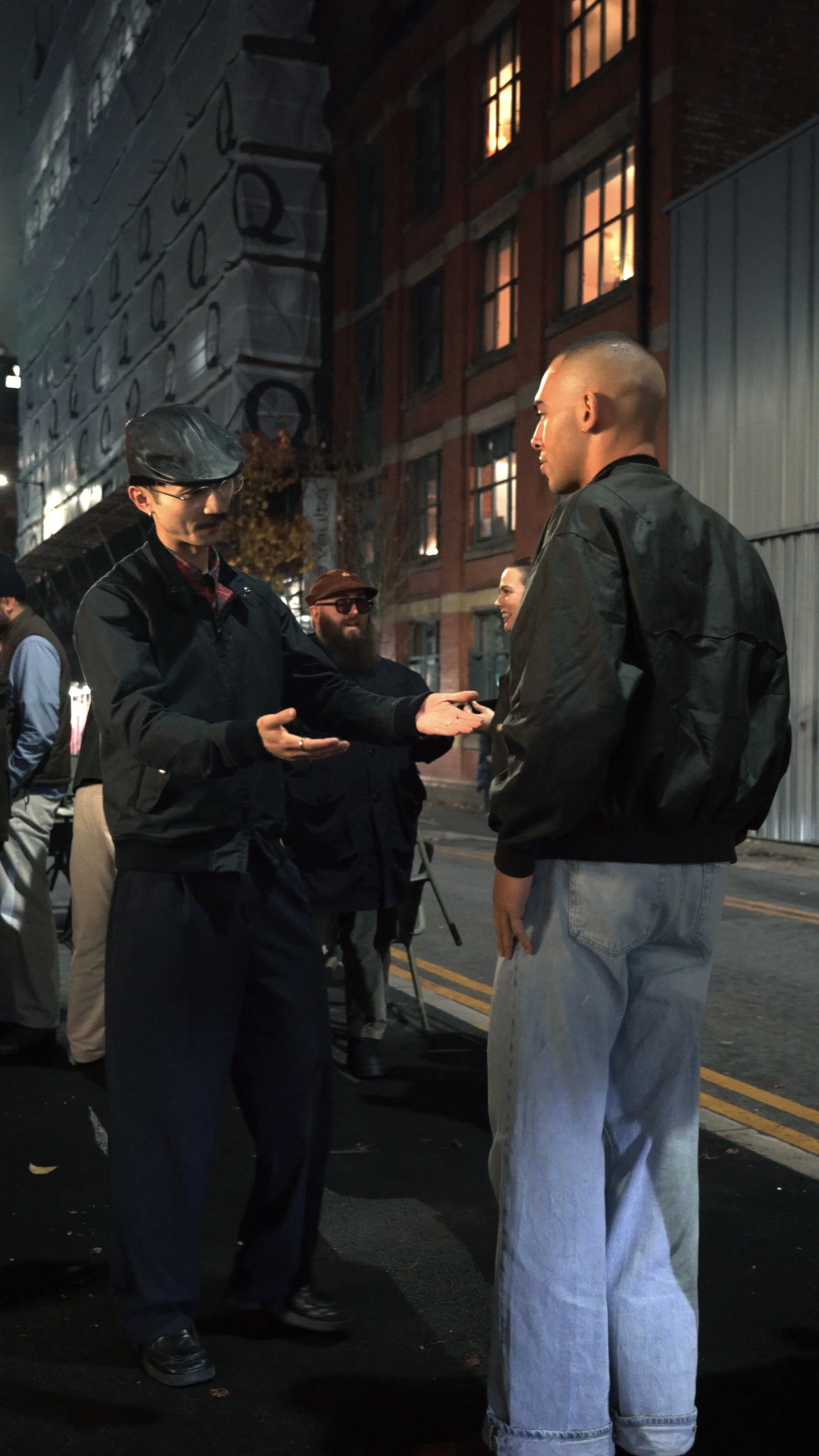 Two people standing face-to-face at night on a city street, engaged in a conversation, with others and buildings illuminated in the background.