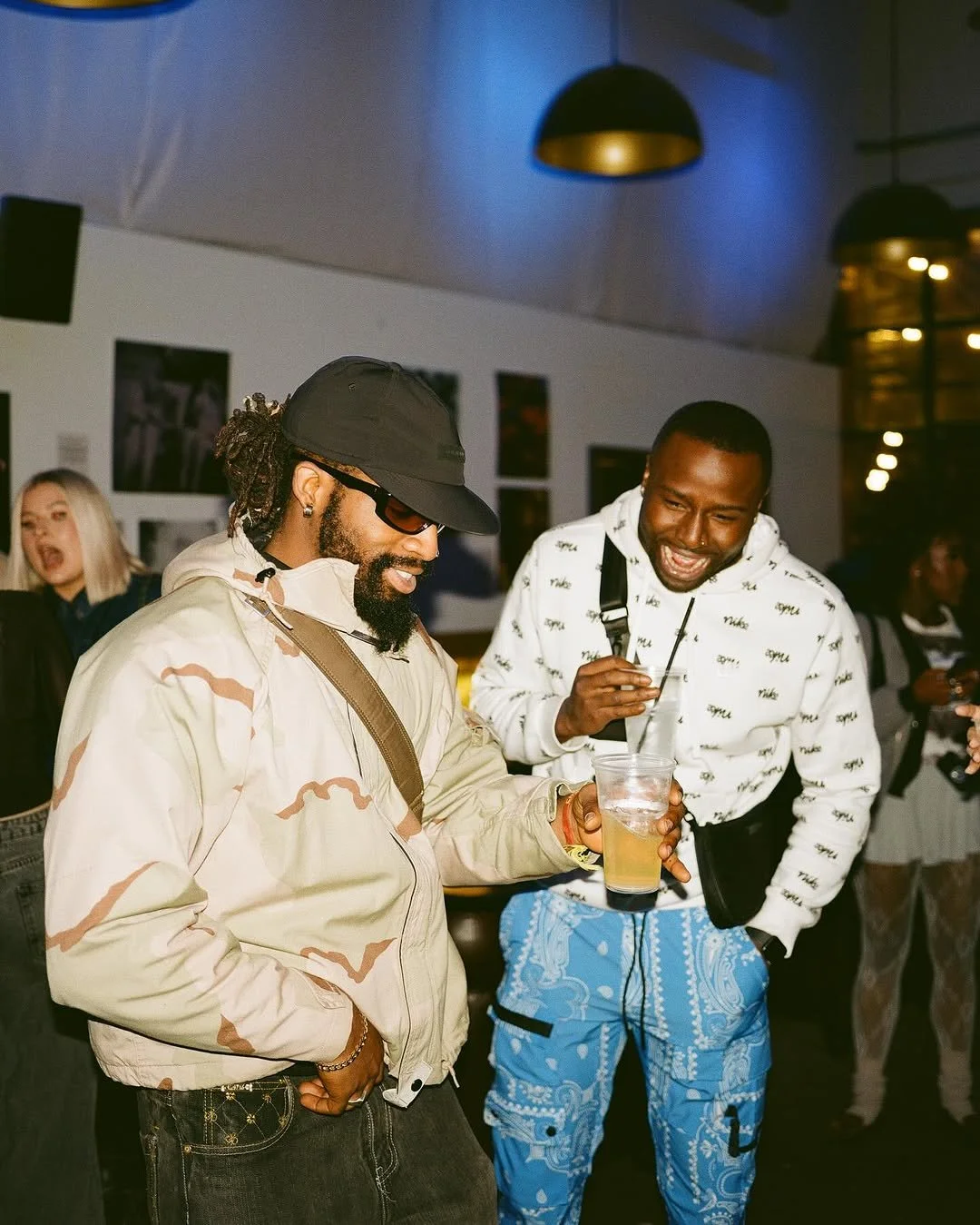 Two men enjoying a drink and laughing in a social setting, with other guests in the background.