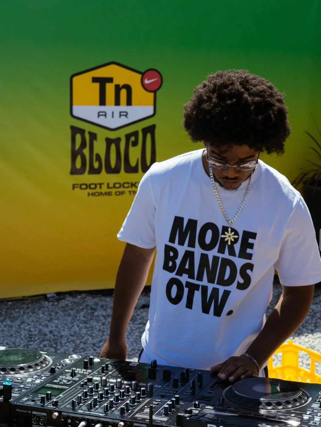 A DJ with curly hair, glasses, wearing a white T-shirt that says 'MORE BANDS. WOT.' and a silver chain, is standing behind a DJ console outdoors, with a green and yellow promotional backdrop for Tn Air Belo logo in the background.