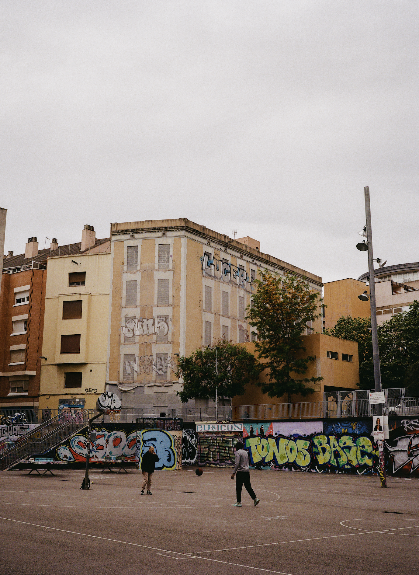 Three people playing basketball on an outdoor court with graffiti-covered walls, trees, and apartment buildings in the background under a cloudy sky.