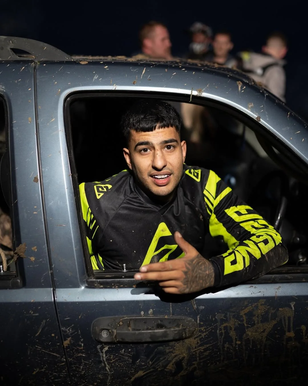 A young man with a black and yellow motocross jersey leaning out of a dirt-covered truck window, gesturing with his hand, with a group of people blurred in the background.