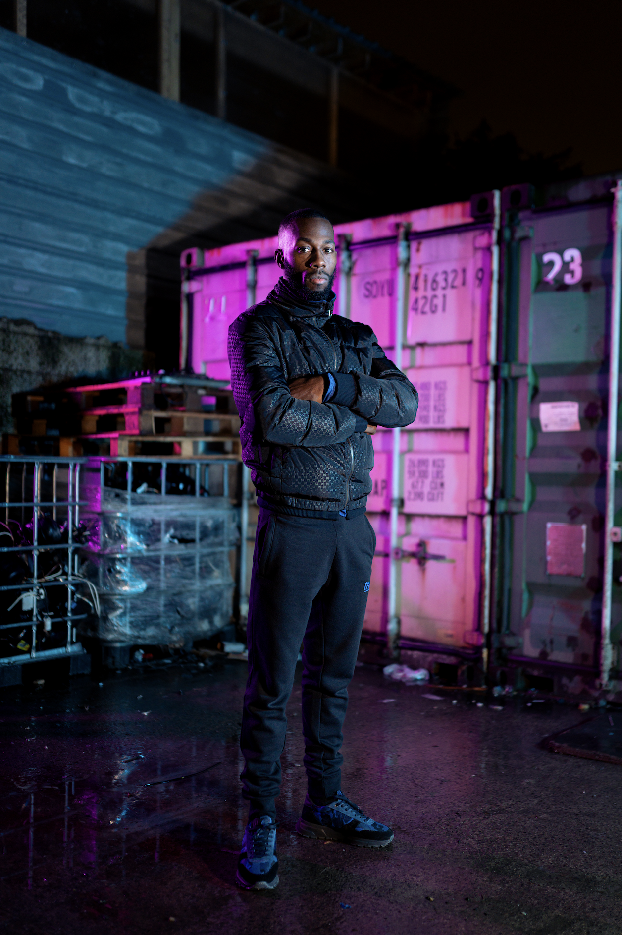 A man stands with arms crossed in front of a colorful, pink-lit shipping container at night, wearing a black jacket and pants.