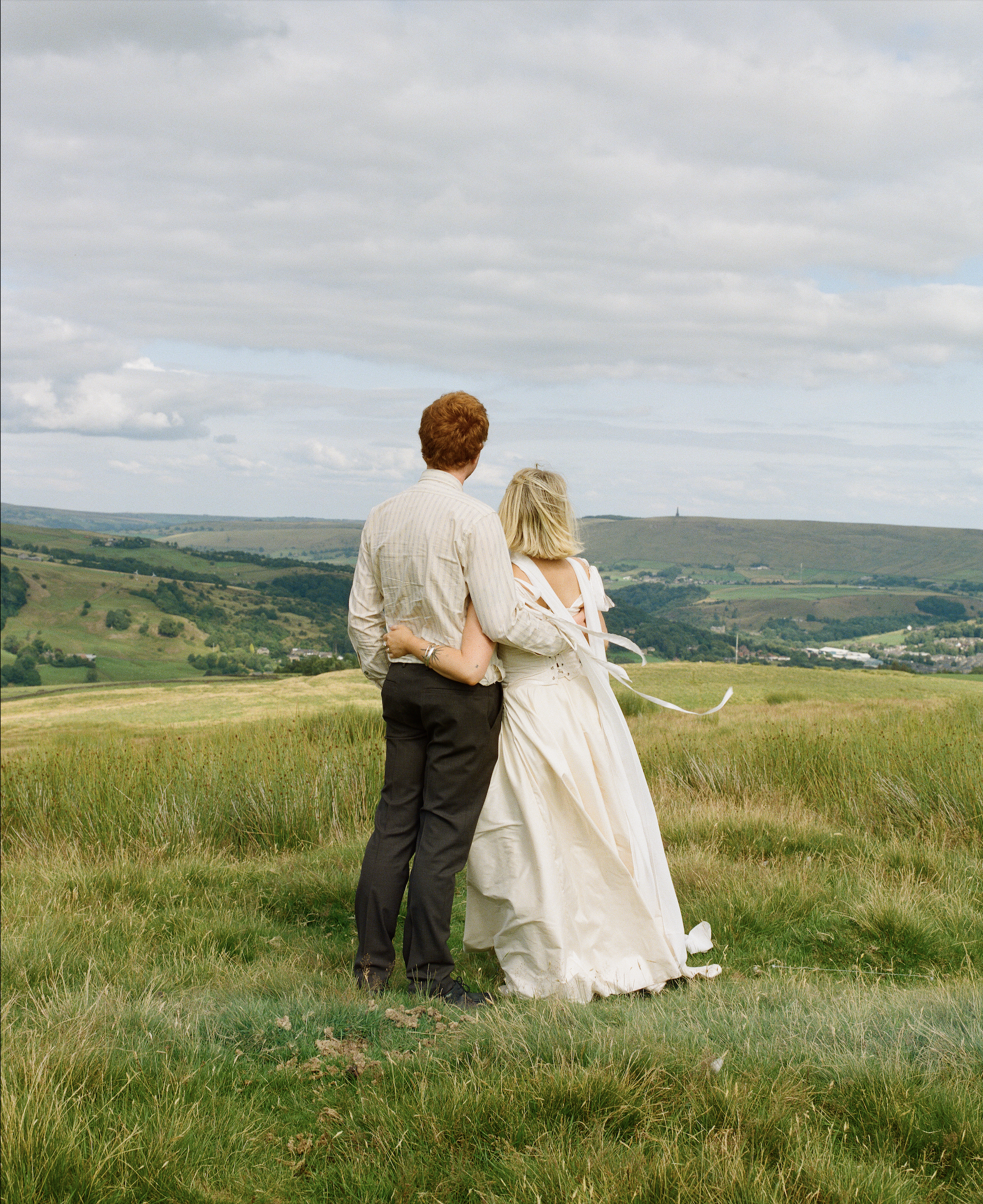 A man and woman stand together on a grassy hilltop, overlooking a scenic countryside landscape with hills and scattered buildings, under a partly cloudy sky. The woman is dressed in a white gown, and the man is in a light shirt and dark trousers.