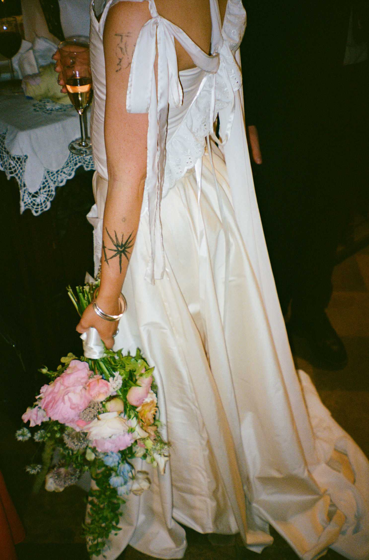 A woman in a white satin dress holding a bouquet of pink and white flowers, with tattoos on her left arm, standing in a room with a table covered with a lace tablecloth and a glass of pink champagne.