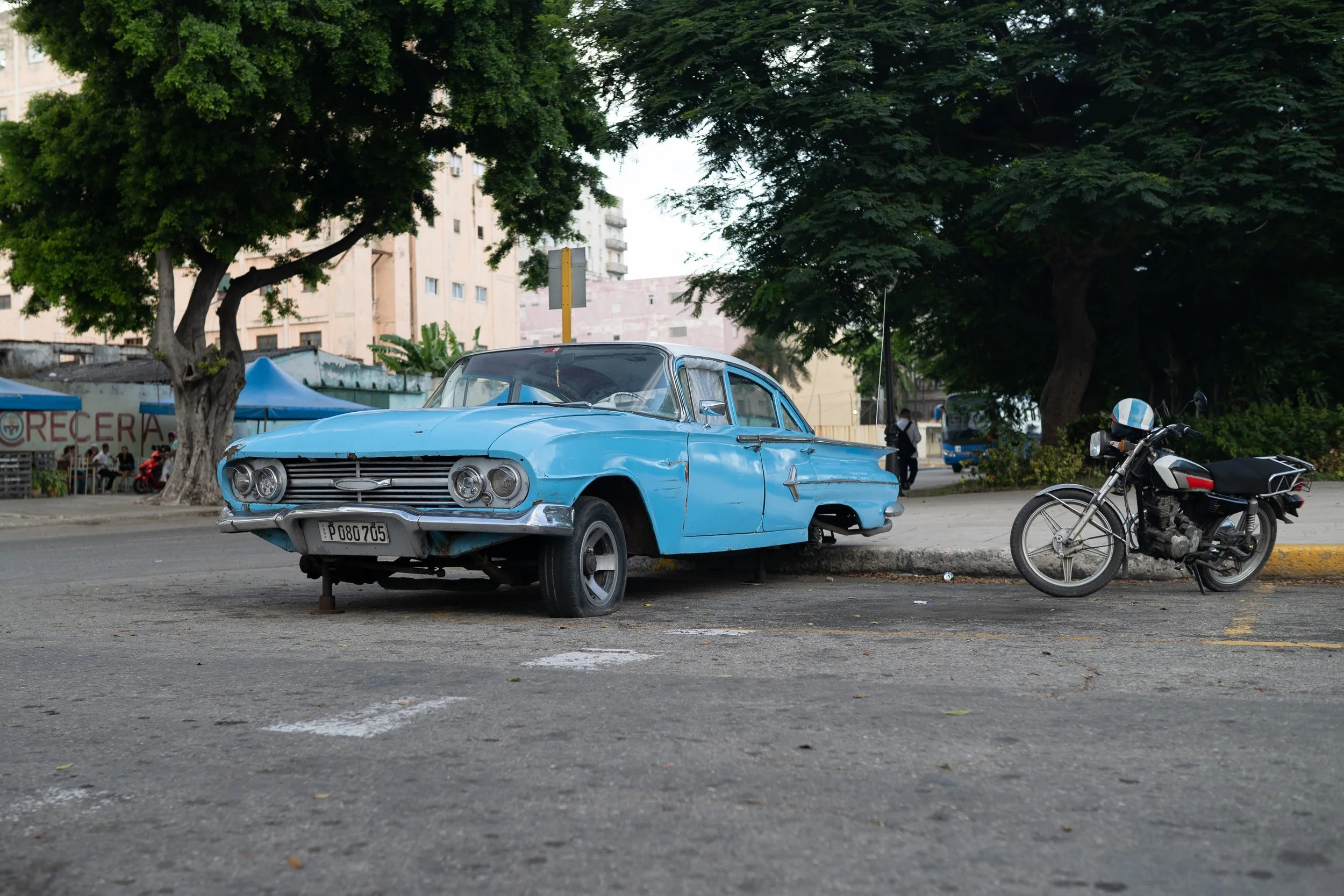 A vintage blue car and a black motorcycle parked on a city street under large green trees, with buildings and a small tent in the background.