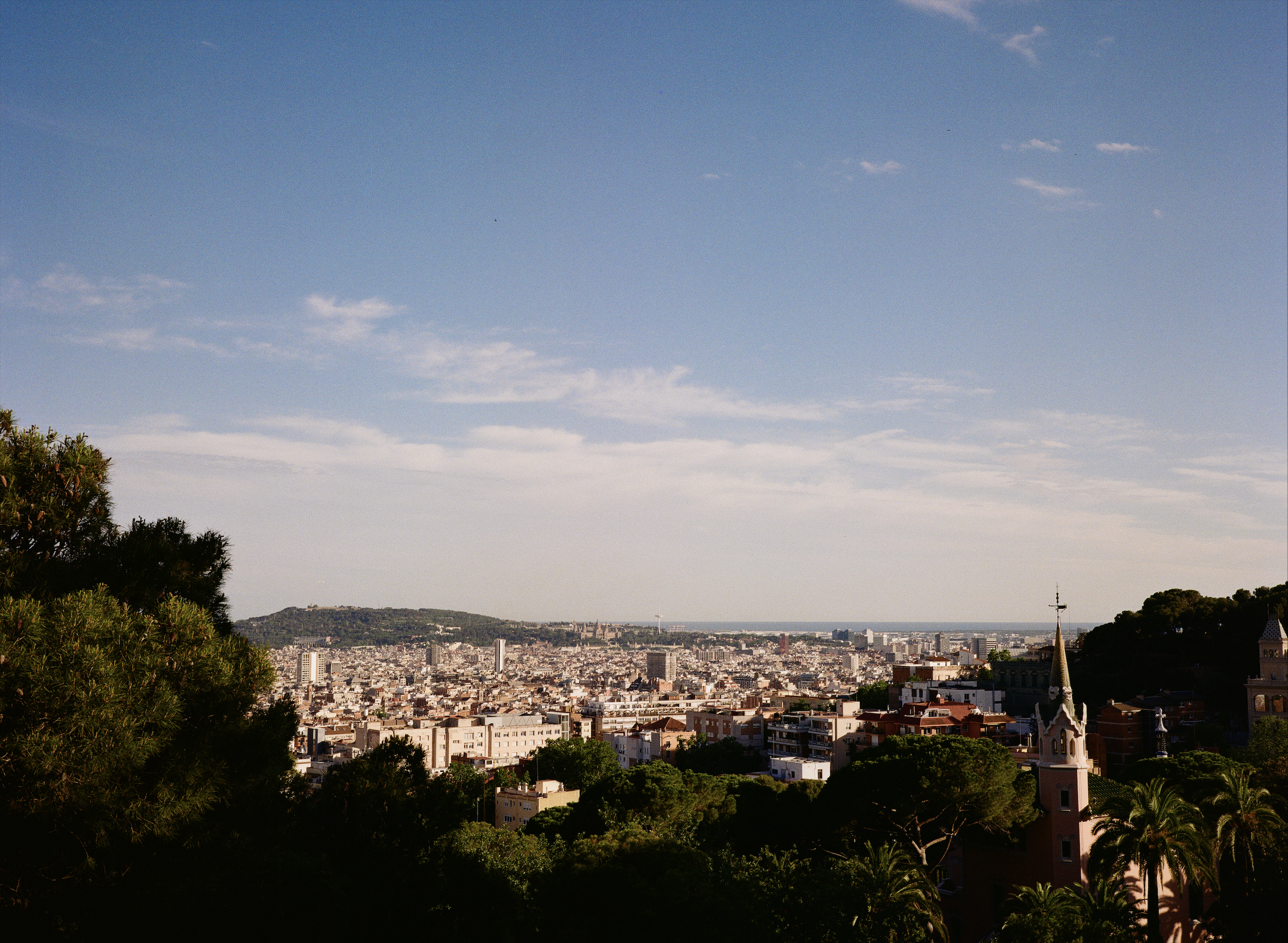 A panoramic view of a cityscape with a mix of modern and historic buildings, greenery, and a church with a tall steeple in the foreground, under a partly cloudy sky.