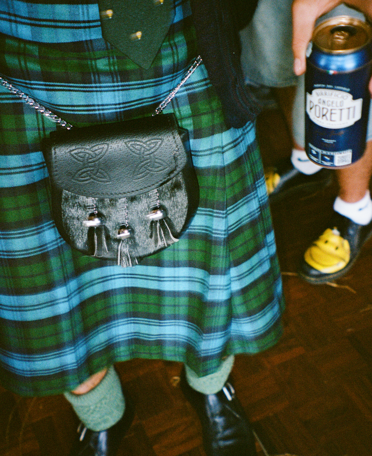 Person wearing a traditional green and blue tartan kilt with green knee-high socks, black shoes, and a black leather sporran, holding a can of beer.