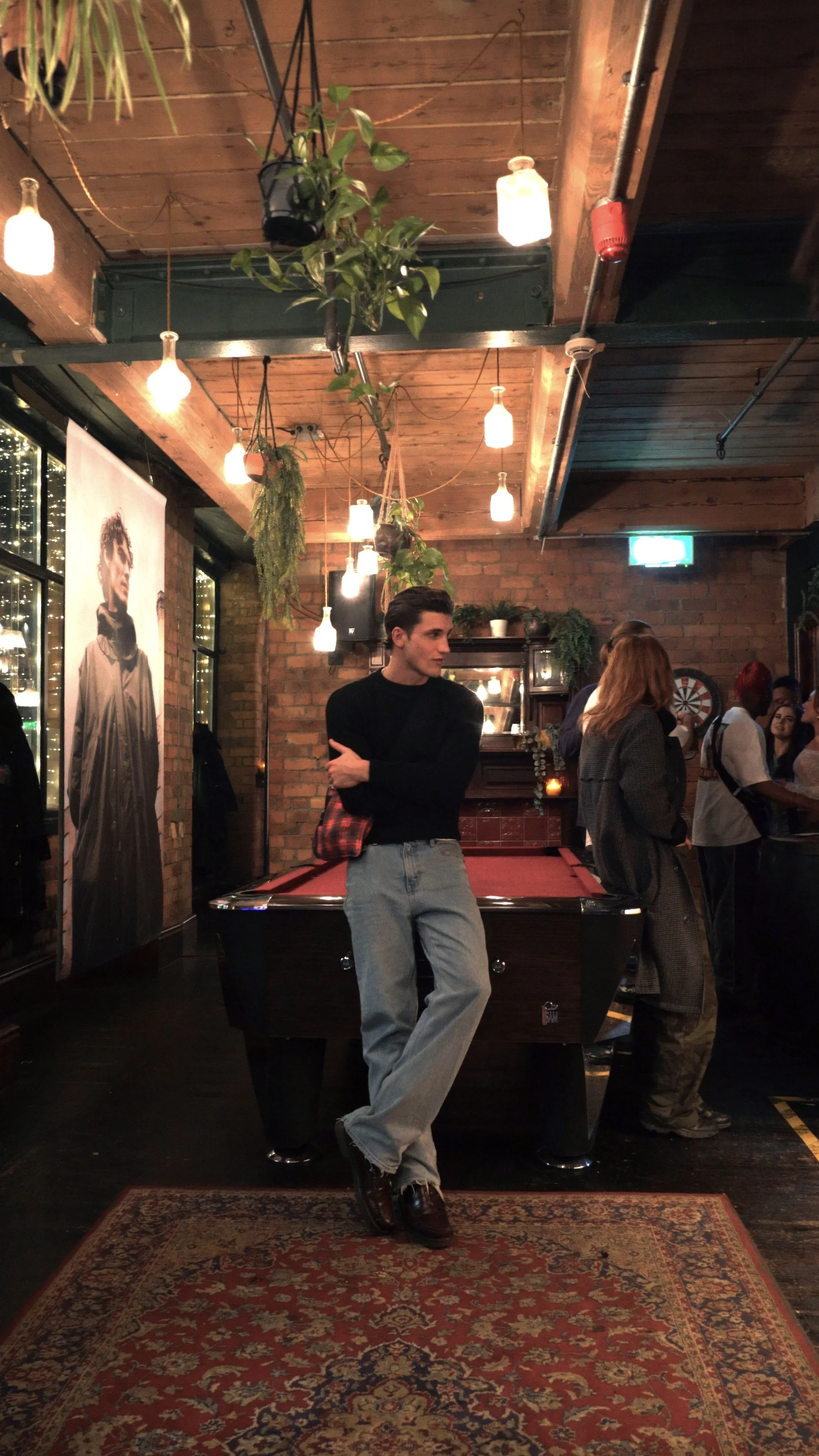 A young man standing with arms crossed in a dimly lit, cozy bar or pub with exposed brick walls, hanging plants, a pool table, and a group of people socializing in the background.