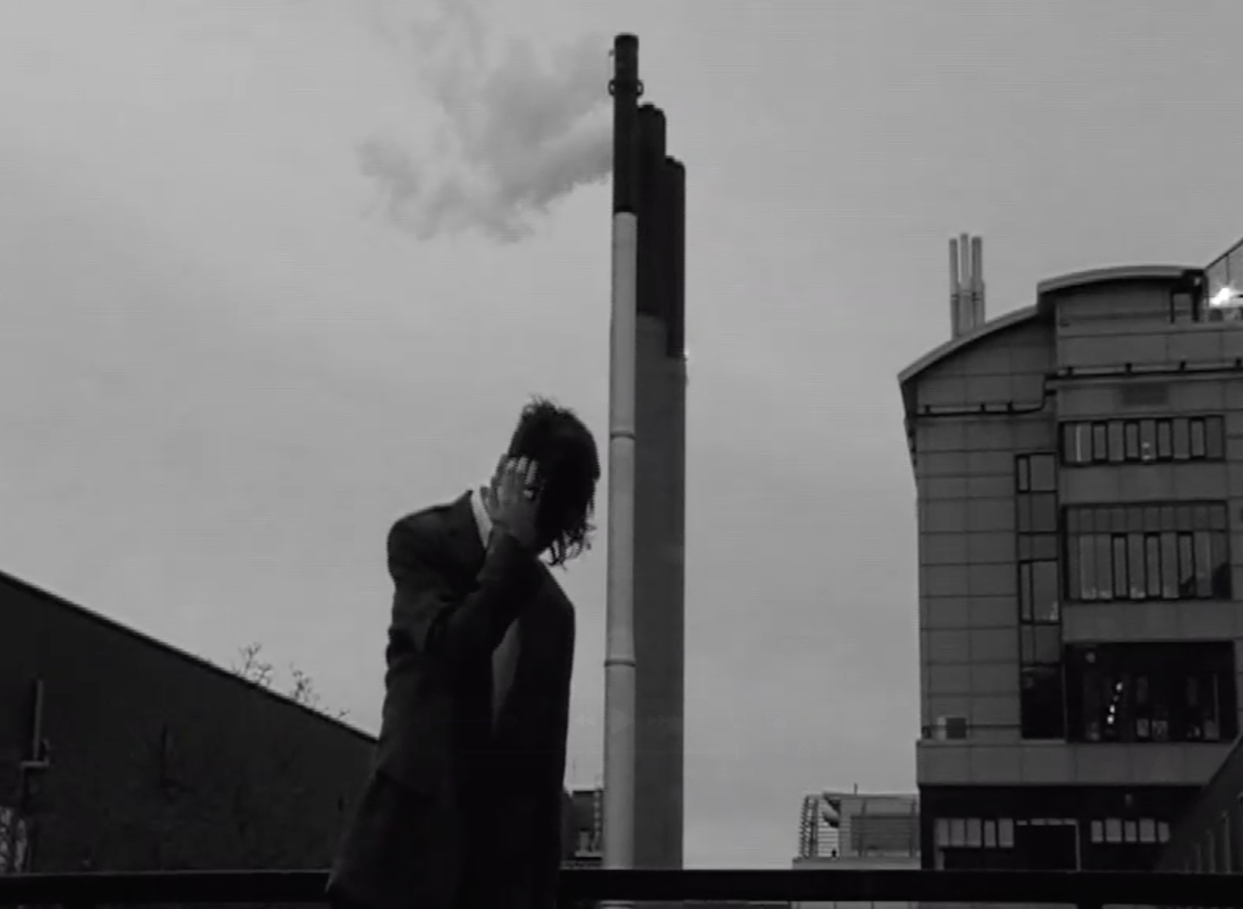 A person with dreadlocks covering their face, wearing a jacket, standing outdoors, covering their face with one hand, with a tall smokestack emitting smoke and modern buildings in the background under a cloudy sky.