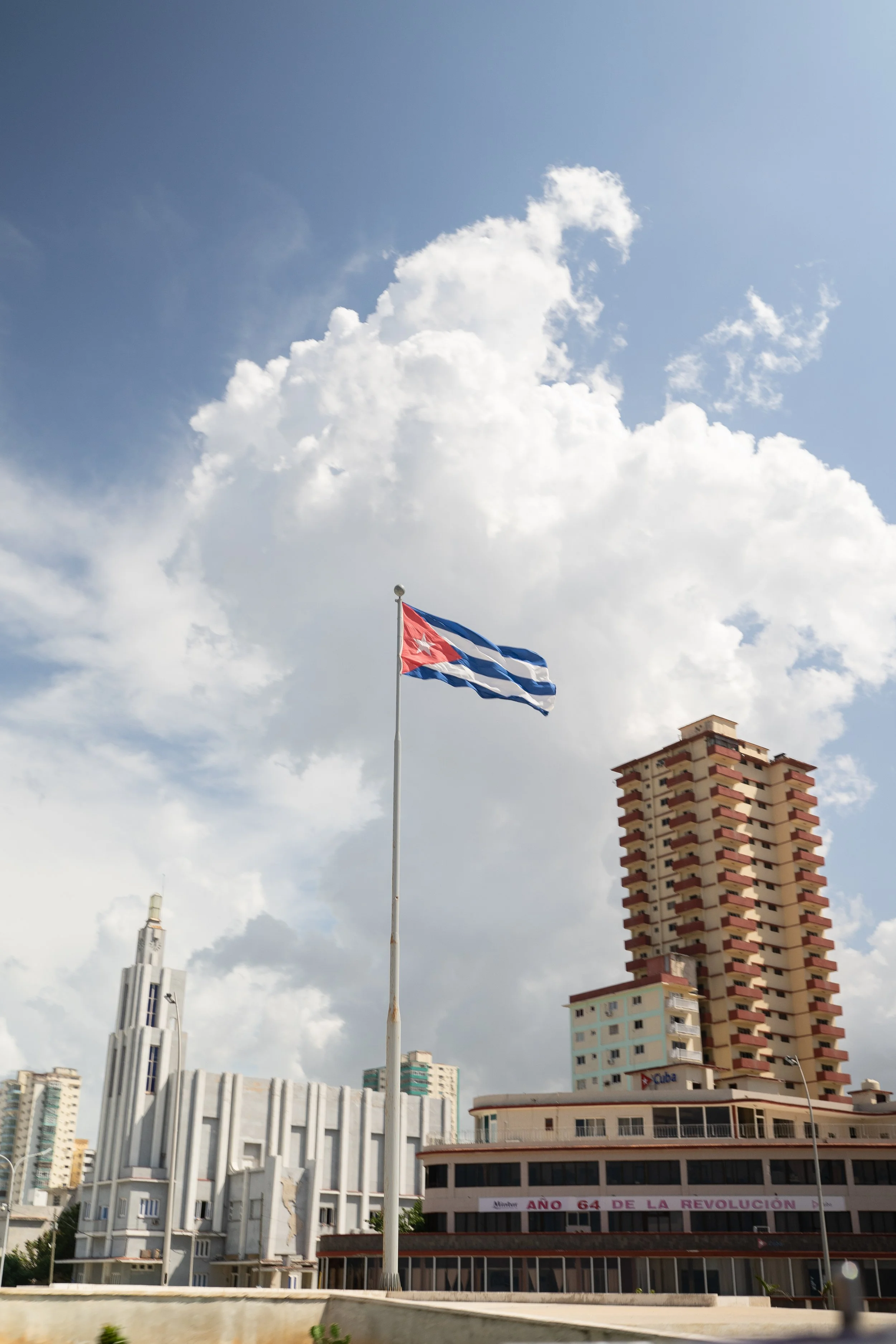 Cuban flag waving in the wind above a cityscape with mid-rise and high-rise buildings under a partly cloudy sky.