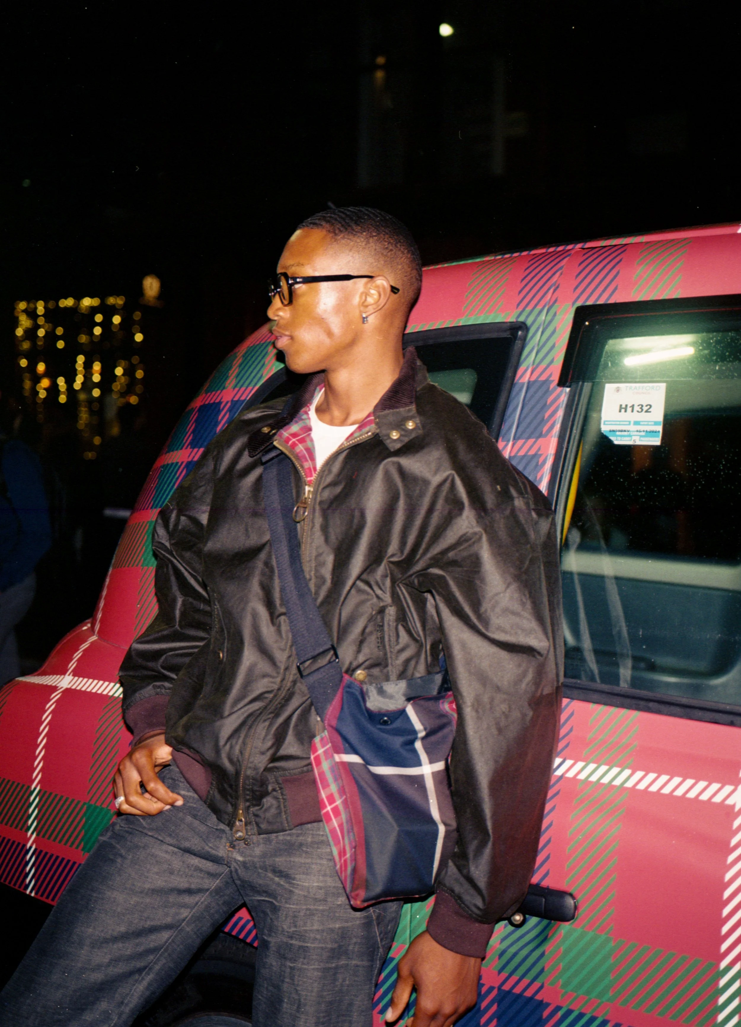 A young man wearing glasses and a black jacket with a plaid lining, leaning against a car with a red, green, and blue tartan pattern at night.