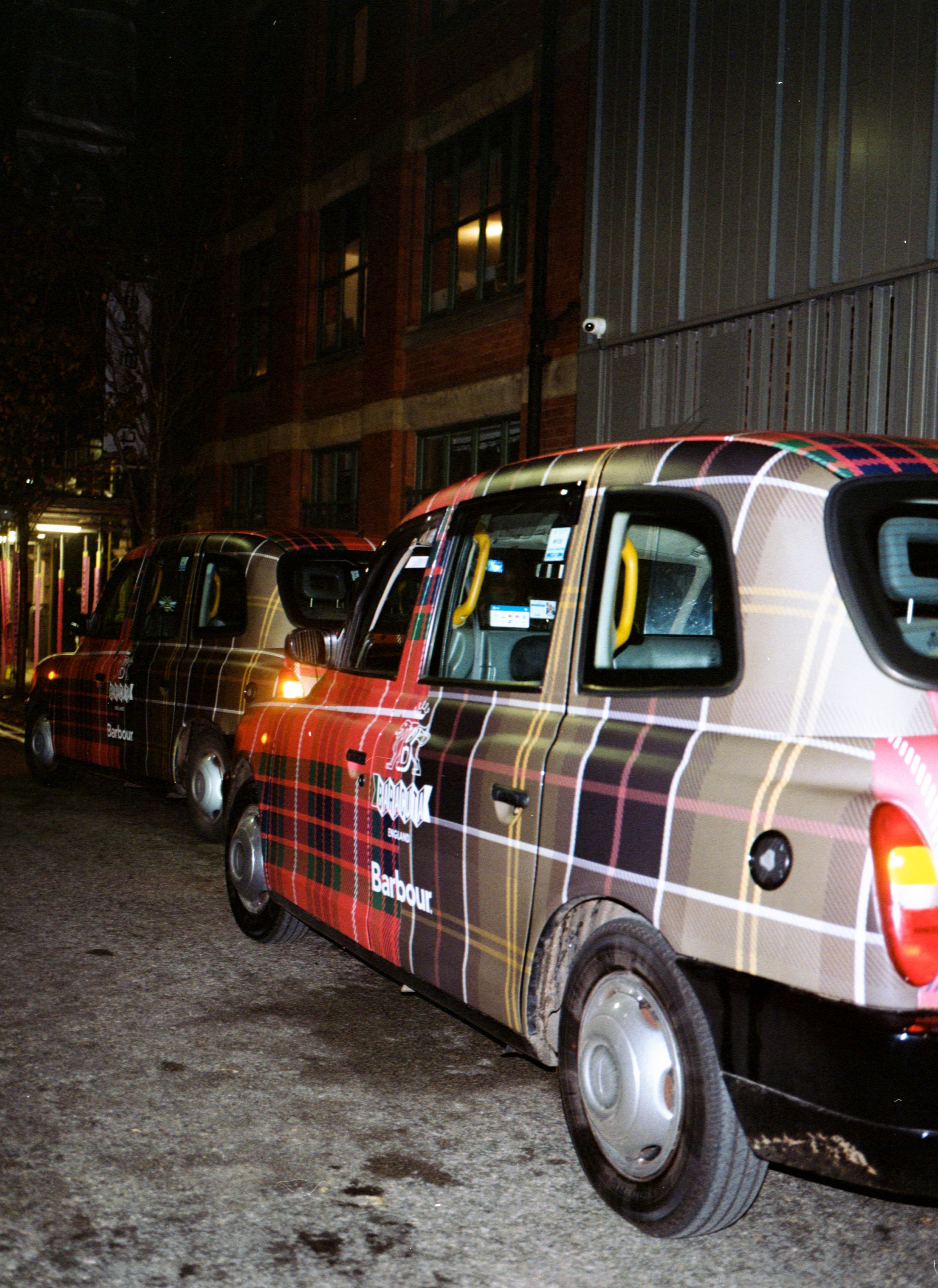Two small cars wrapped in tartan plaid patterns, parked on a city street at night, with a brick building in the background.