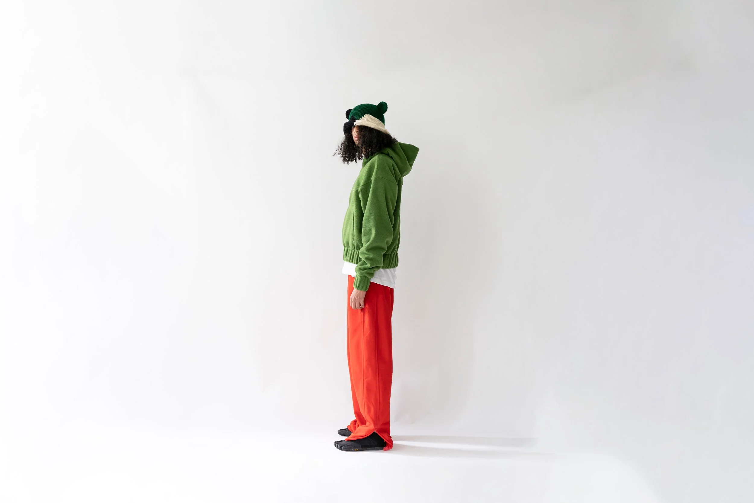 A person standing sideways against a plain white background, wearing a green hoodie, red pants, black shoes, and a knit hat with green and beige bear ears.