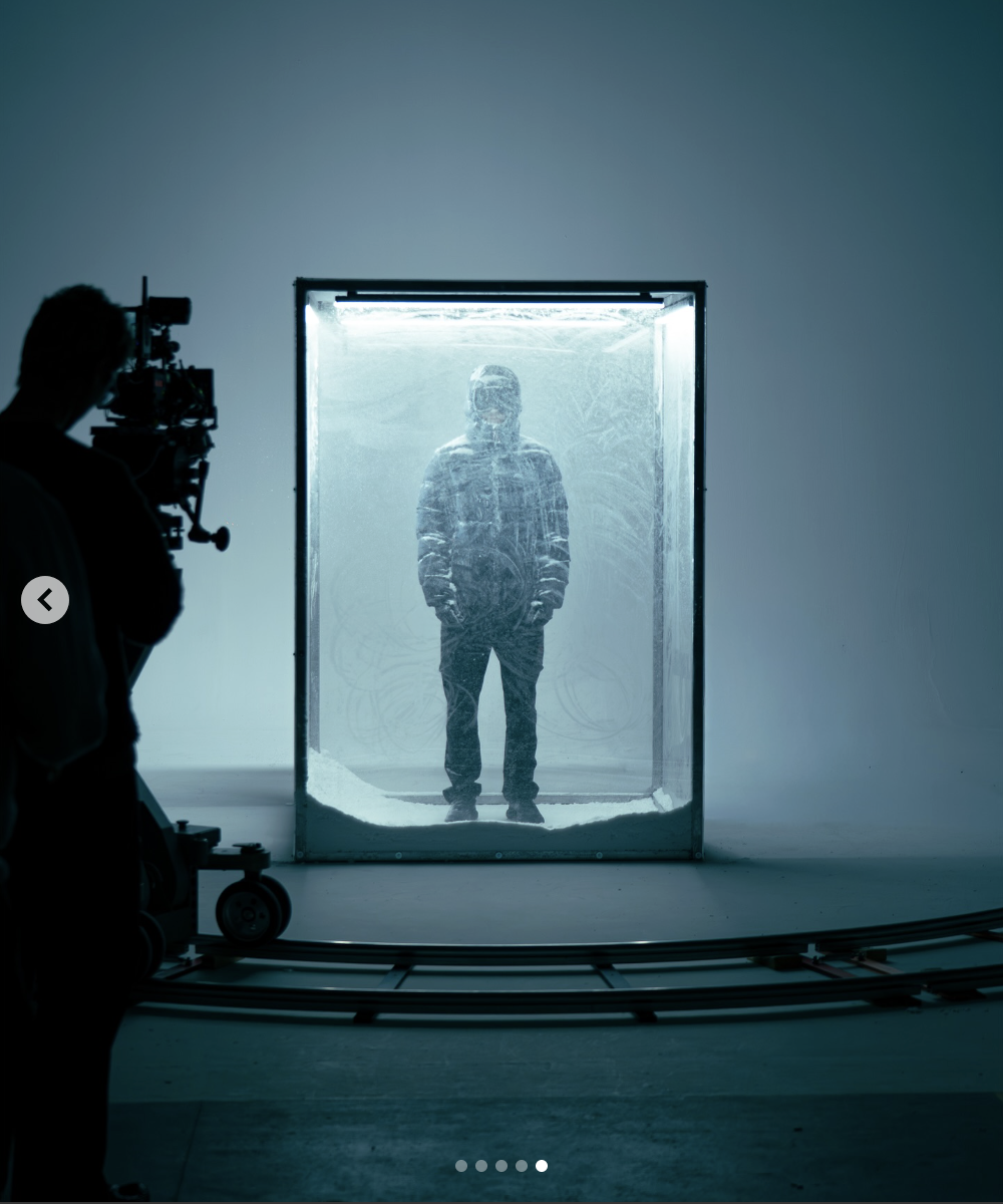 Person standing inside a clear glass tank filled with what appears to be sand, with a photographer taking a picture of him. The scene is set in a minimalist room with plain walls and a circular track on the floor.