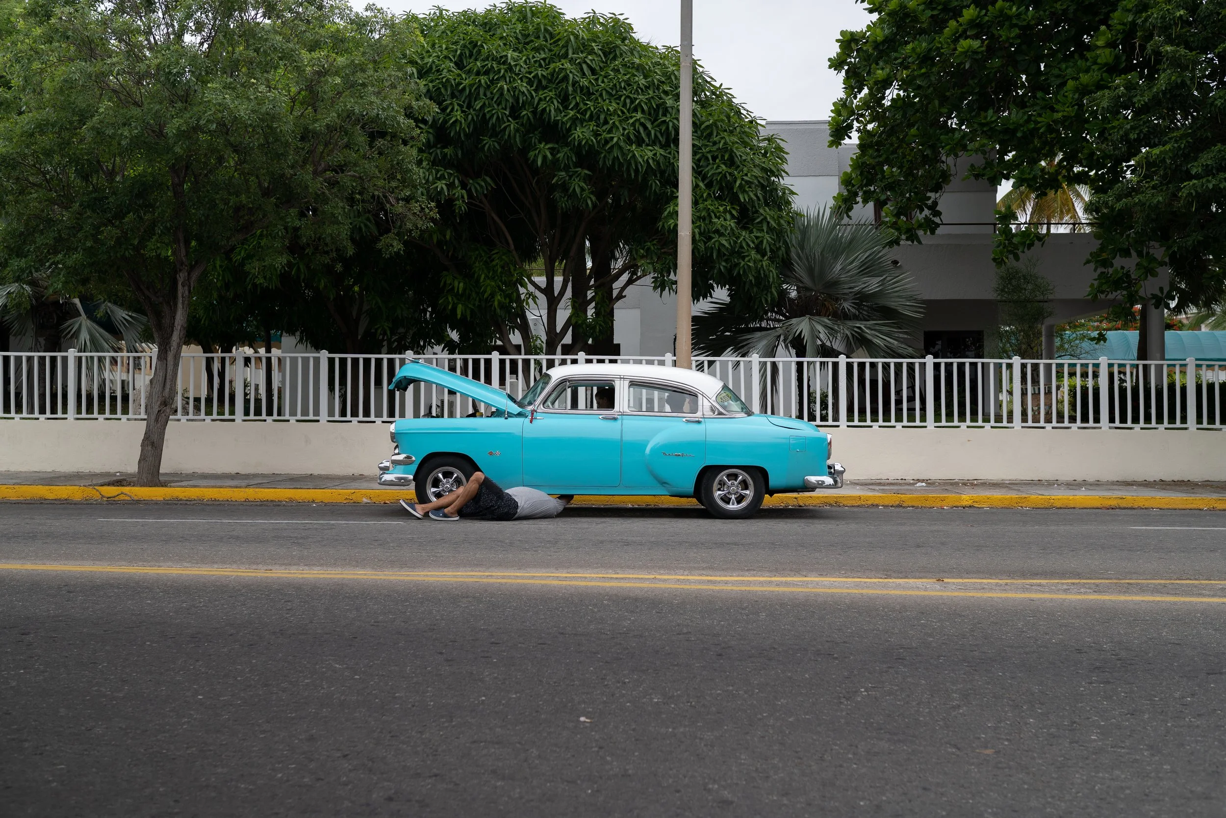 A man lying under the front of a vintage blue car with the hood open on the side of the street, with trees and a white fence in the background.