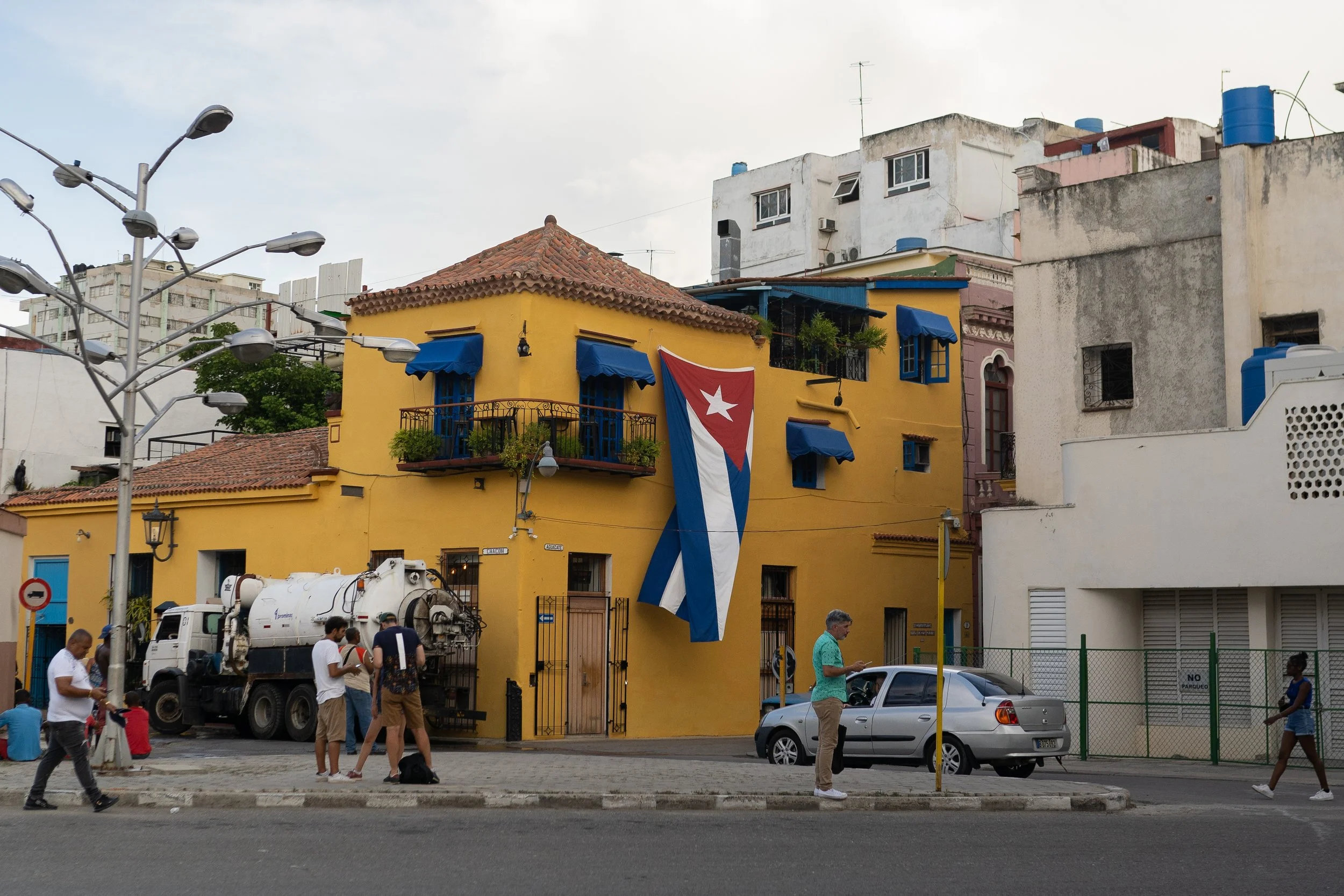 Colorful yellow building with blue window awnings and a large Cuban flag hanging from the side. People are walking on the street, and a truck is parked in front of the building.