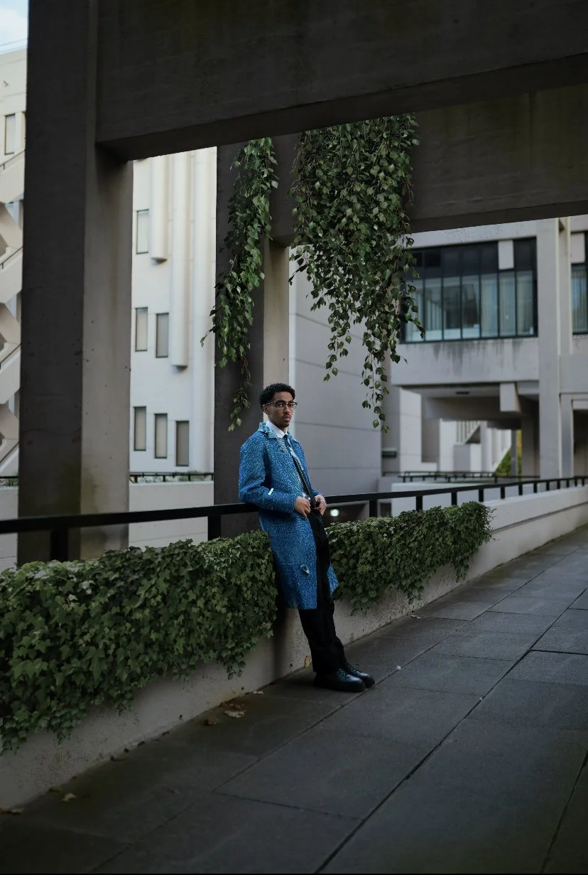 A man in a blue coat leaning against a concrete wall with greenery in an urban setting.