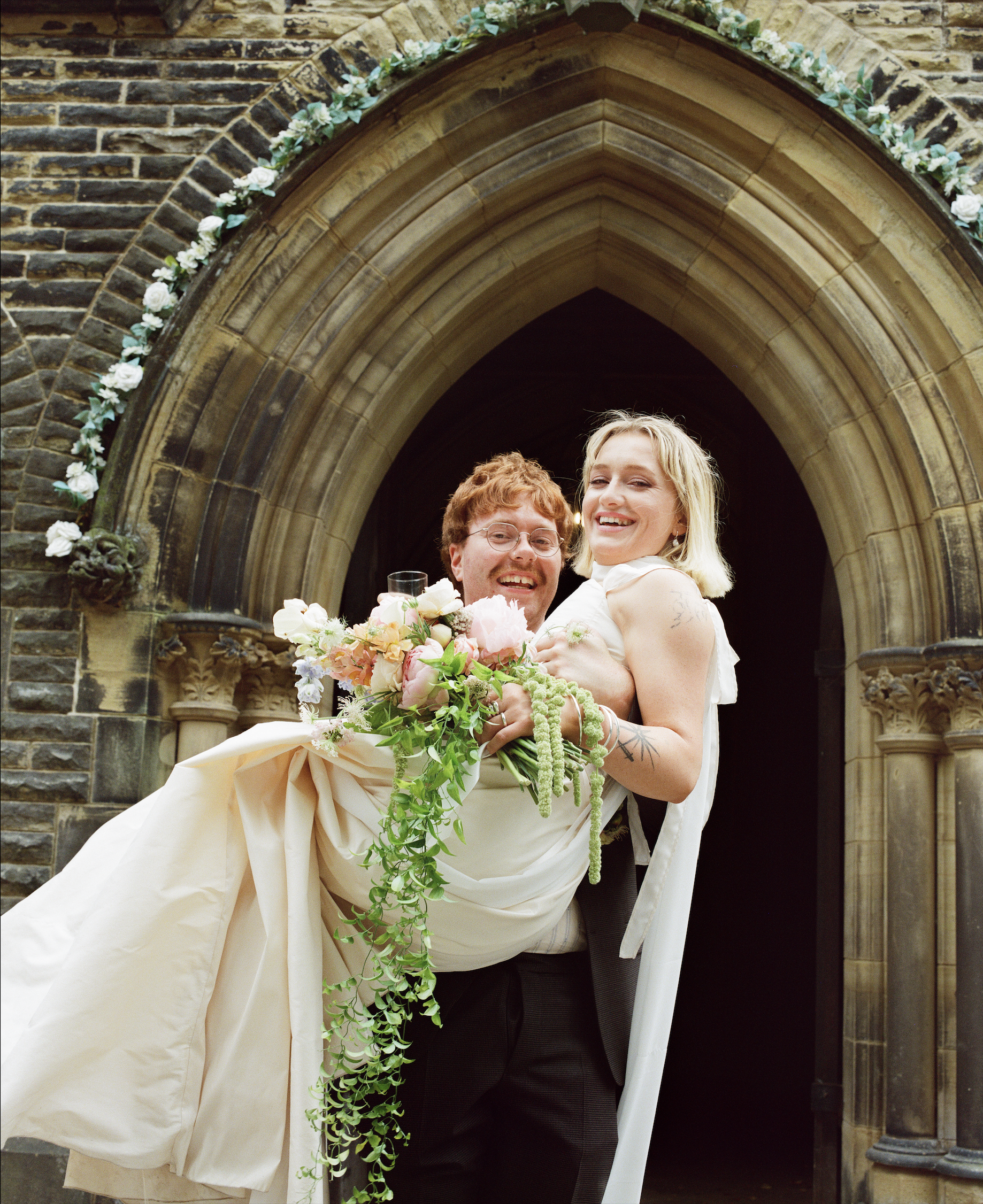 A man in a suit carrying a woman in a wedding dress holding a bouquet of flowers, standing in front of a stone church entrance decorated with white flowers for a wedding photoshoot.
