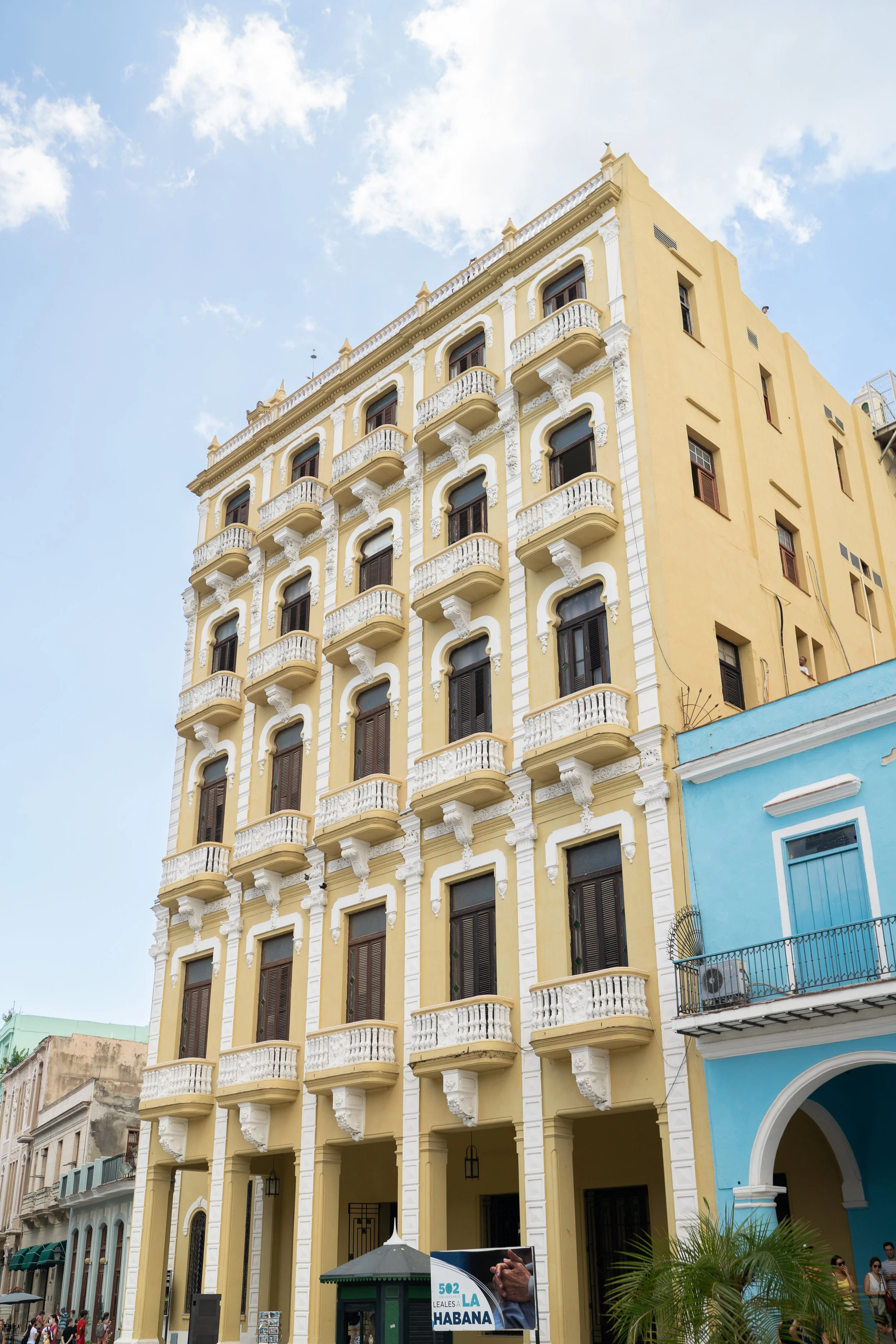 Tall, yellow building with ornate white balconies and dark windows in a city street scene under a blue sky.